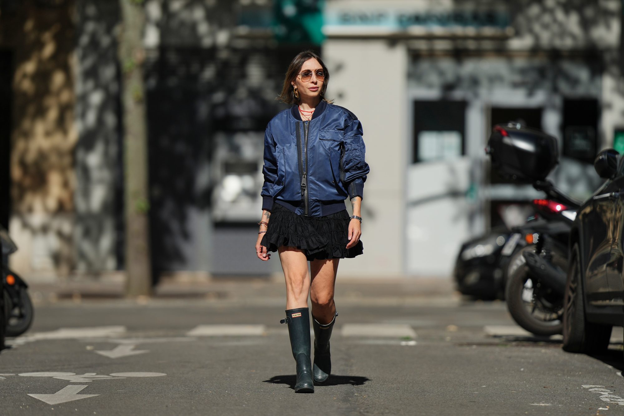 PARIS, FRANCE - SEPTEMBER 06: Emy Venturini wears straight light brown hair with a middle part, brown gradient sunglasses by Prada, gold hoop earrings by Rabanne, layered necklaces, multiple bracelets, a navy blue bomber jacket by Burberry, a short ruffled black dress by Zara Studio, and dark green rubber rain boots by Hunter, during a street style fashion photo session, on September 06, 2025 in Paris, France. (Photo by Edward Berthelot/Getty Images)