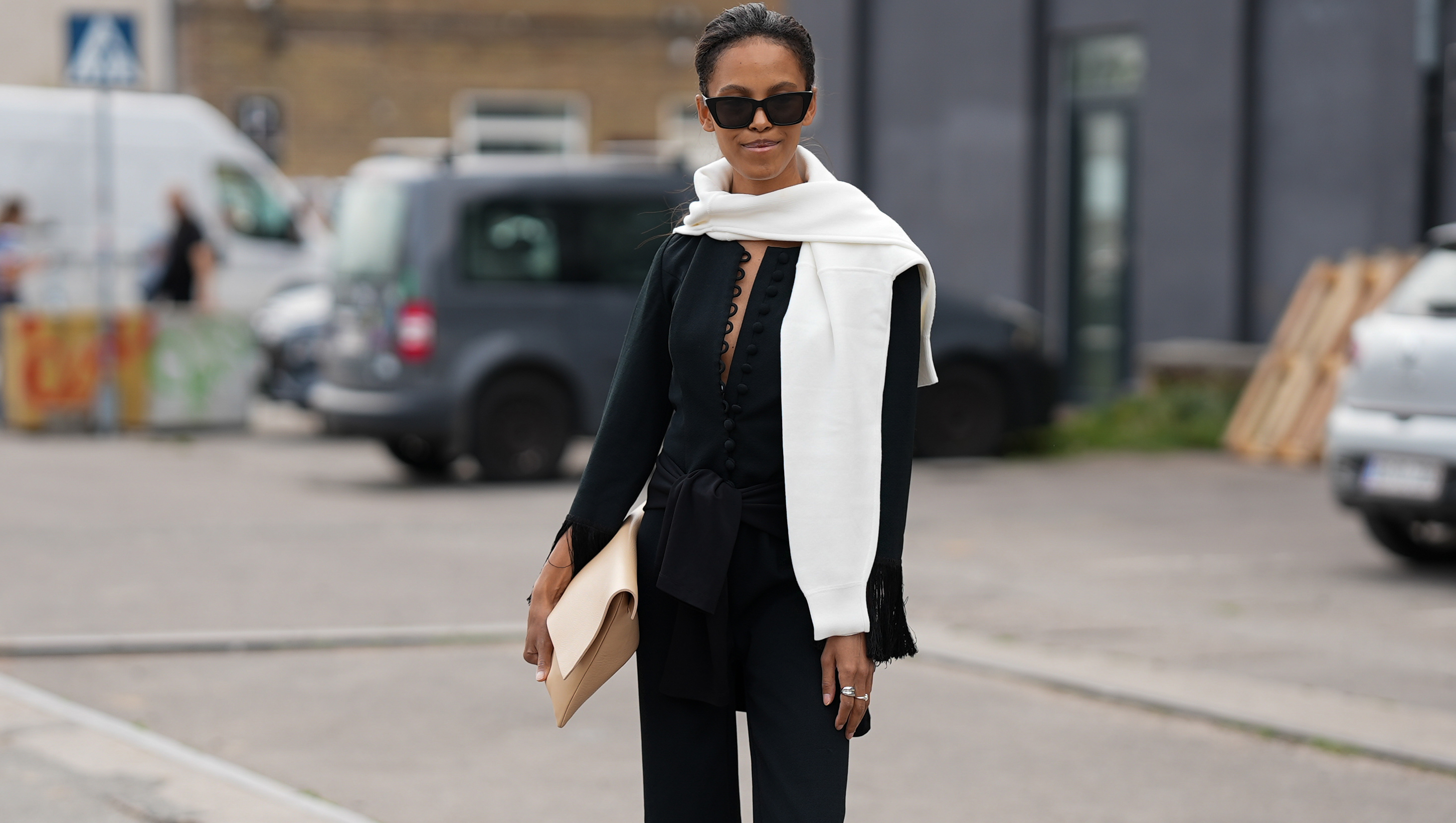 COPENHAGEN, DENMARK - AUGUST 7: Suzann&radic;&dagger; Gembege is seen wearing a white knit sweater draped over the shoulders; a black blouse with a deep V-neck big button details and fringe-accented sleeves; a matching black fringe-hem trousers; black flip flops; a black cat-eye sunglasses; a beige oversized clutch; silver rings; her black hair is styled straight in a ponytail outside the Marimekko show during Copenhagen Fashion Week day four on August 7, 2025 in Copenhagen, Denmark. (Photo by Moritz Scholz/Getty Images)