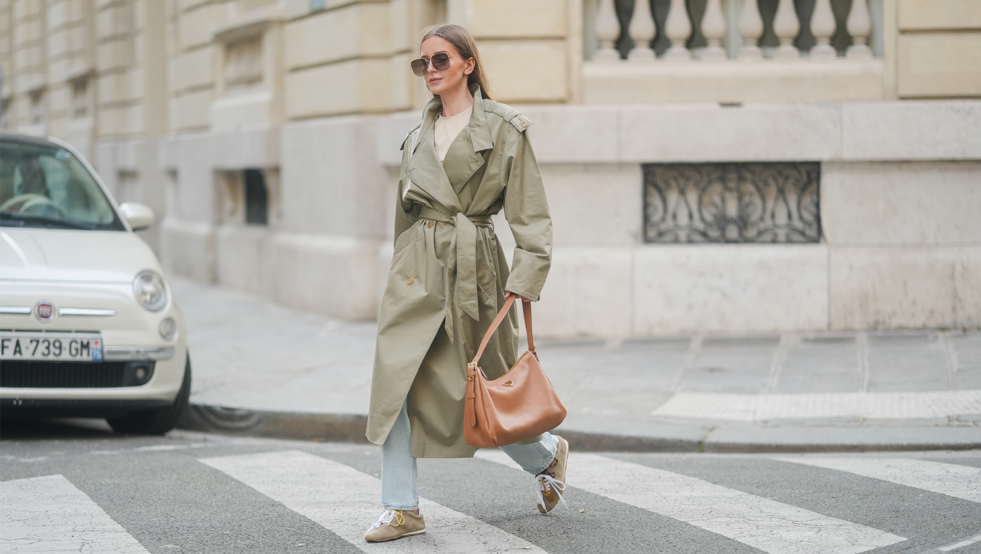 Diane Batoukina wears her long straight brown hair down with a center part, oversized square Dior sunglasses, a khaki belted trench coat by Sandro layered over a beige T-shirt by Zara, light-wash straight-leg jeans by Anine Bing, beige suede Miu Miu sneakers with mismatched laces and tassel details, and carries a large tan leather shoulder bag by Prada, walking across a pedestrian crossing, during a street style fashion photo session on March 25, 2025 in Paris, France.