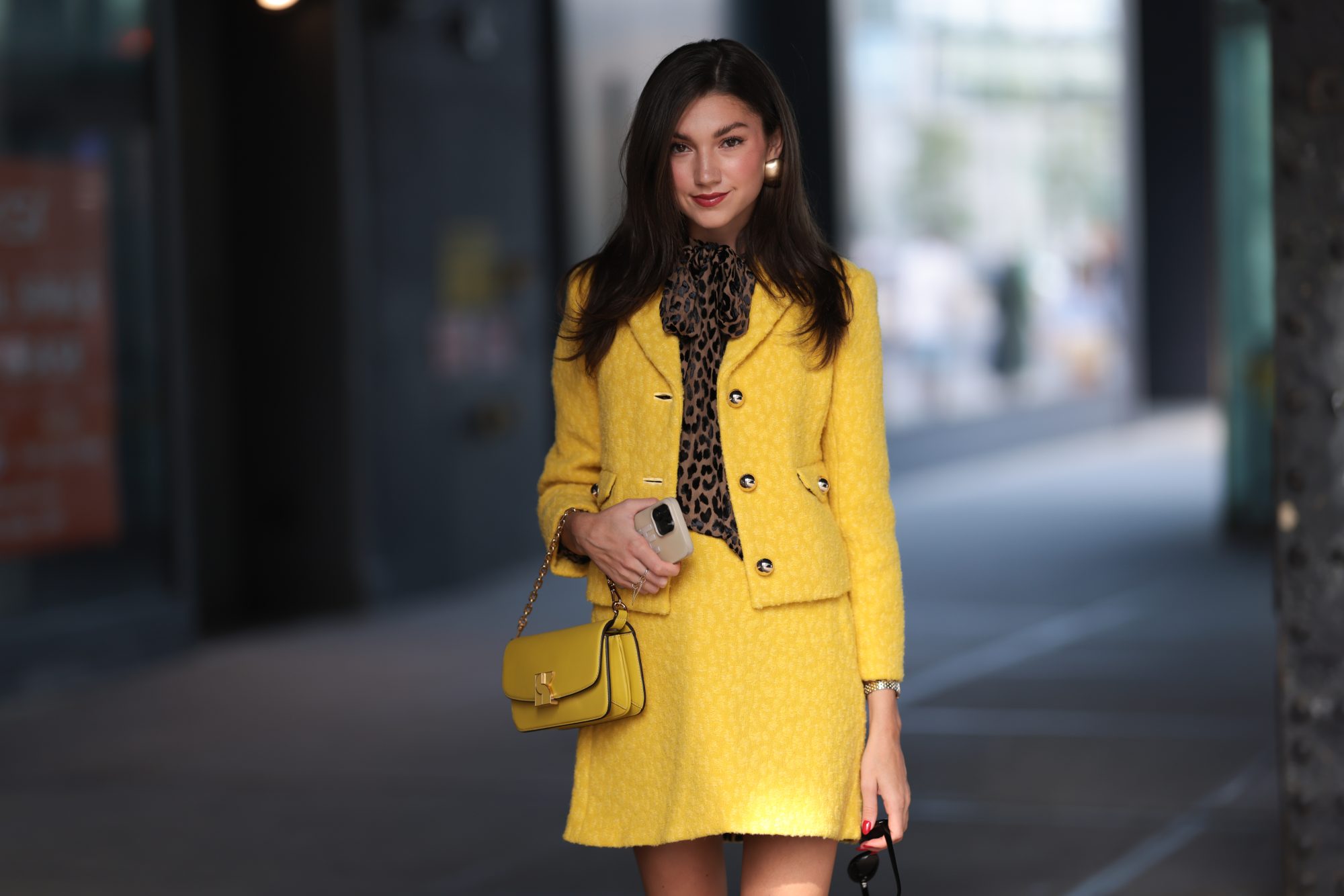 NEW YORK, NEW YORK - SEPTEMBER 08: A Fashion Week guest is seen wearing black sunglasses, golden earrings, a yellow matching two-piece from Kate Spade consisting of a jacket with golden buttons and a mini skirt, underneath a brown/black top with animal print and a bow, a silver/golden watch, a yellow leather bag from Kate Spade and pumps with ankle strap and animal print from Kate Spade outside before Kate Spade presentation on September 08, 2023 in New York City. (Photo by Jeremy Moeller/Getty Images)