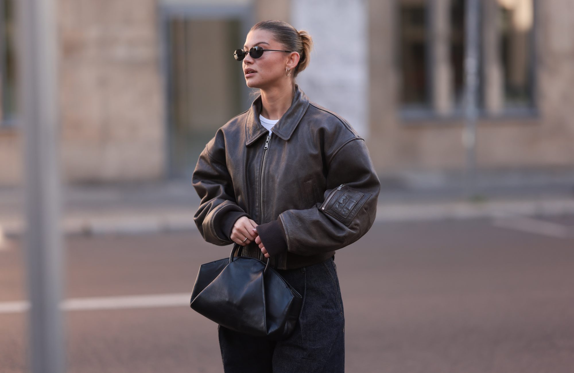 BERLIN, GERMANY - OCTOBER 12: Sophia Geiss is seen wearing oval rimless black sunglasses from Vehla Eyewear, two different-sized gold hoop earrings, a silver nose ring, a cropped dark brown leather jacket with wide sleeves and ribbed cotton cuffs from Meotine, a white top underneath, wide-legged dark vintage jeans with orange decorative seams, a silver ring, and a black leather bag with round handles from COS on October 12, 2024 in Berlin, Germany. (Photo by Jeremy Moeller/Getty Images)
