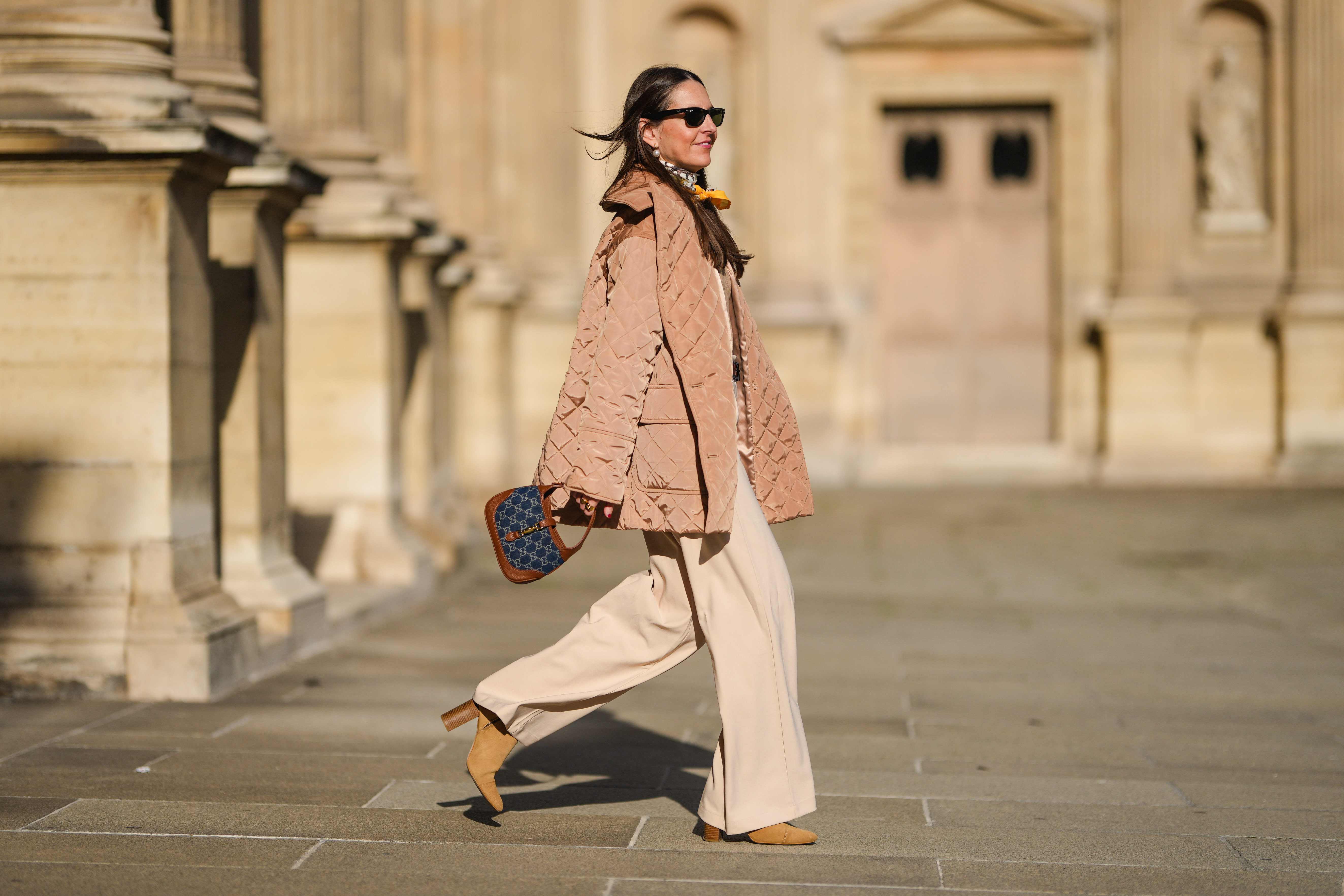 Alba Garavito Torre wears pearls earrings, black wayfarer sunglasses from Ray Ban, a beige quilted oversized jacket from Munthe, a white and yellow silk scarf tie with classic knot from Lollys Laundry, a brown / beige braided wool pullover from Polo Ralph Lauren, a brown shiny leather belt, high waist beige palazzo pants from Bimba y Lola, a blue denim monogram and brown shiny leather Jackie 1961 Mini bag handbag from Gucci, yellow mustard suede block heels ankle boots, gold bracelets, gold rings, during a street style fashion photo session, on November 12, 2021 in Paris, France.