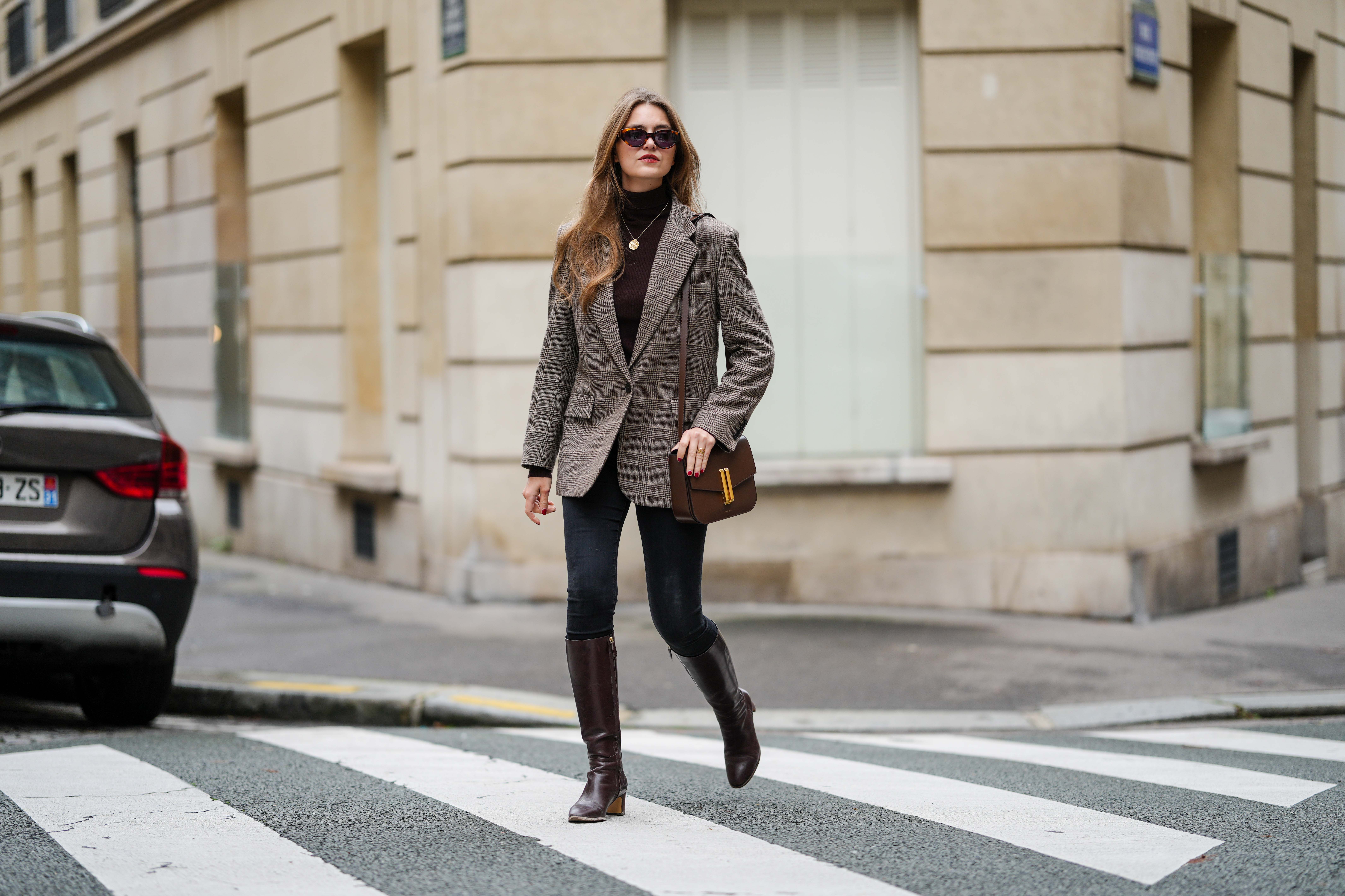 Segolene Hyppolite wears brown Jimmy Fairly sunglasses, gold necklace, grey checked Maje blazer jacket, a dark brown Maje turtleneck pullover, black denim jeans / pants, dark brown leather Rivecour boots, a brown leather Demellier London bag, during a street style fashion photo session, on October 12, 2024 in Paris, France.