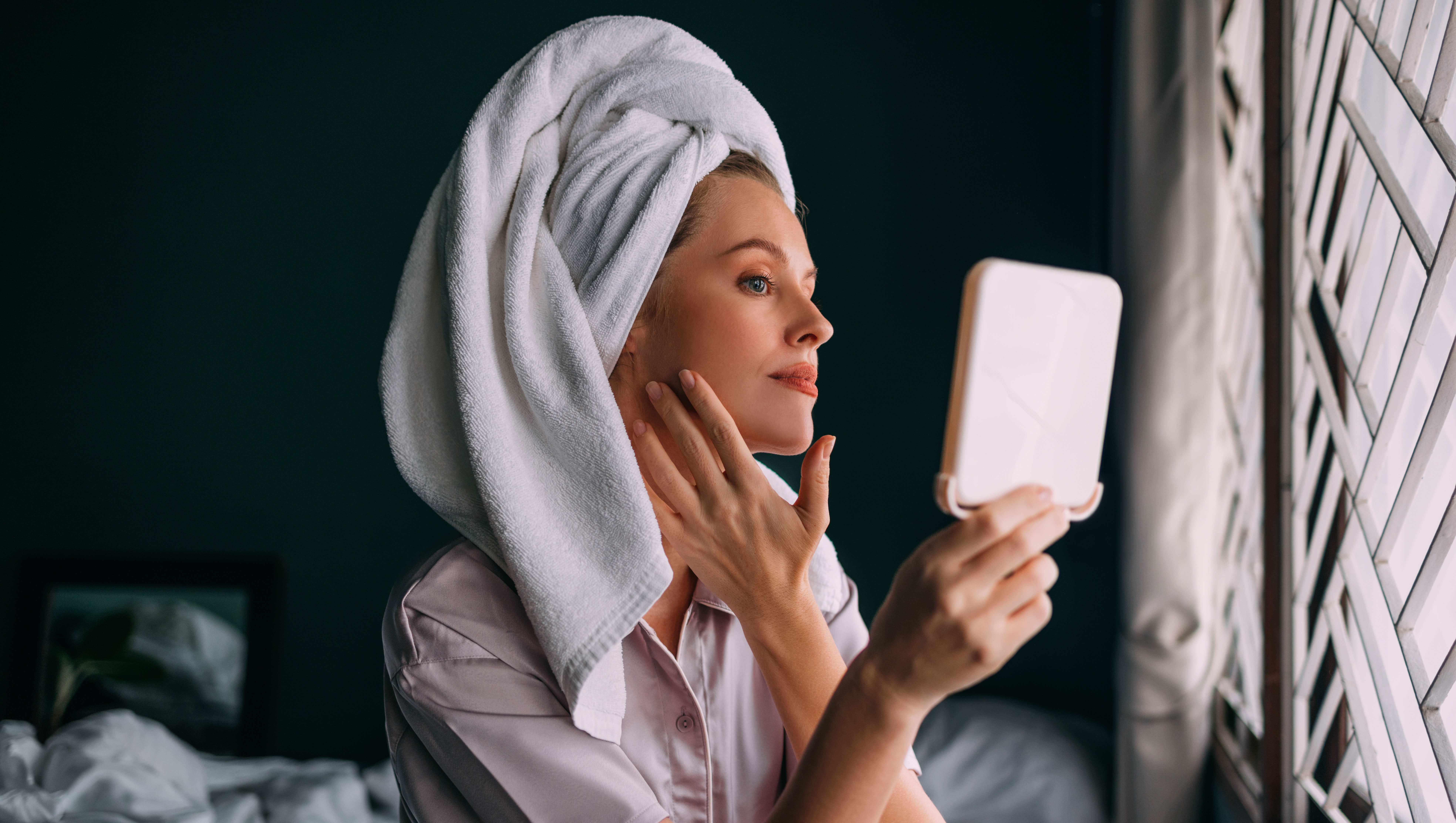 A pensive Caucasian female with her hair wrapped up in a towel keeping her face clean and hydrated using different cosmetic products. She is sitting on the bed in her bedroom. (skincare concept)