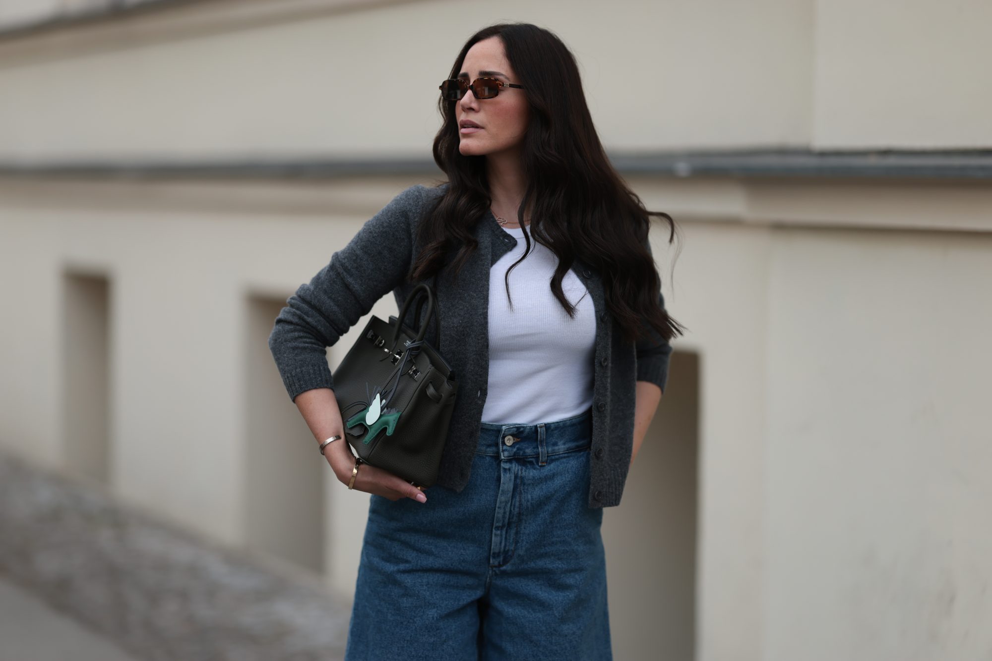 BERLIN, GERMANY - JUNE 27: Raffaela Taschlizki is seen wearing rectangular Havana-framed sunglasses with brown lenses and thin temples from Miu Miu; a gold necklace with a diamond-studded letter pendant; an unbuttoned grey wool cardigan, paired with a white tank top underneath, both from Zara; denim shorts with white logo embroidery at the front from Loewe; a rectangular black leather “Birkin” bag with a horse-shaped “Rodeo” bag charm, both from Hermès; and two “Love” bracelets in yellow gold and white gold from Cartier on June 27, 2025 in Berlin, Germany. (Photo by Jeremy Moeller/Getty Images)
