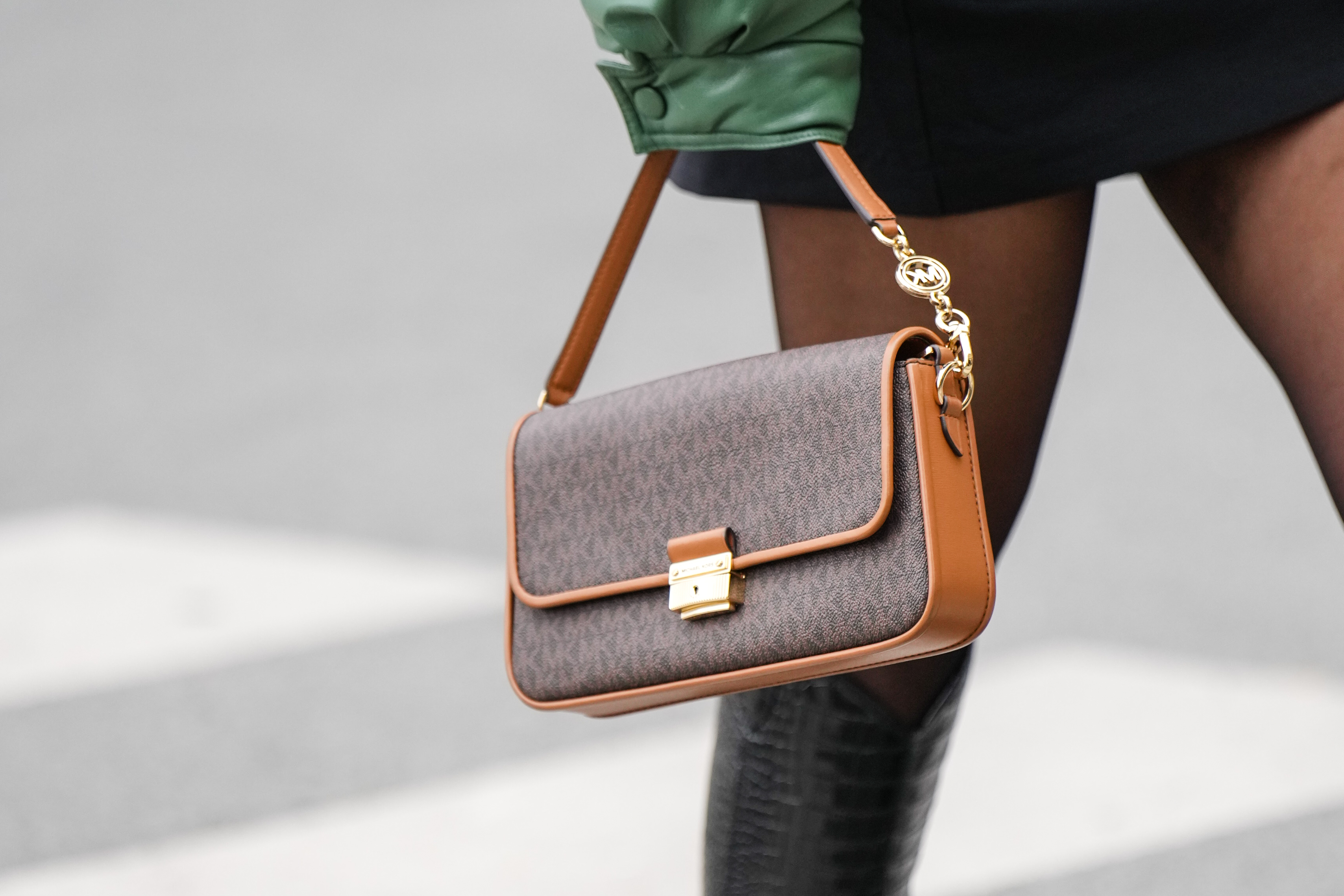 A guest wears a black turtleneck short tube dress, a khaki shiny leather oversized bomber coat, black tights, a brown monogram print pattern in coated canvas handbag from Mickael Kors, black shiny varnished leather crocodile print pattern pointed knees boots, outside Gauchere, during Paris Fashion Week - Womenswear F/W 2022-2023, on March 03, 2022 in Paris, France.