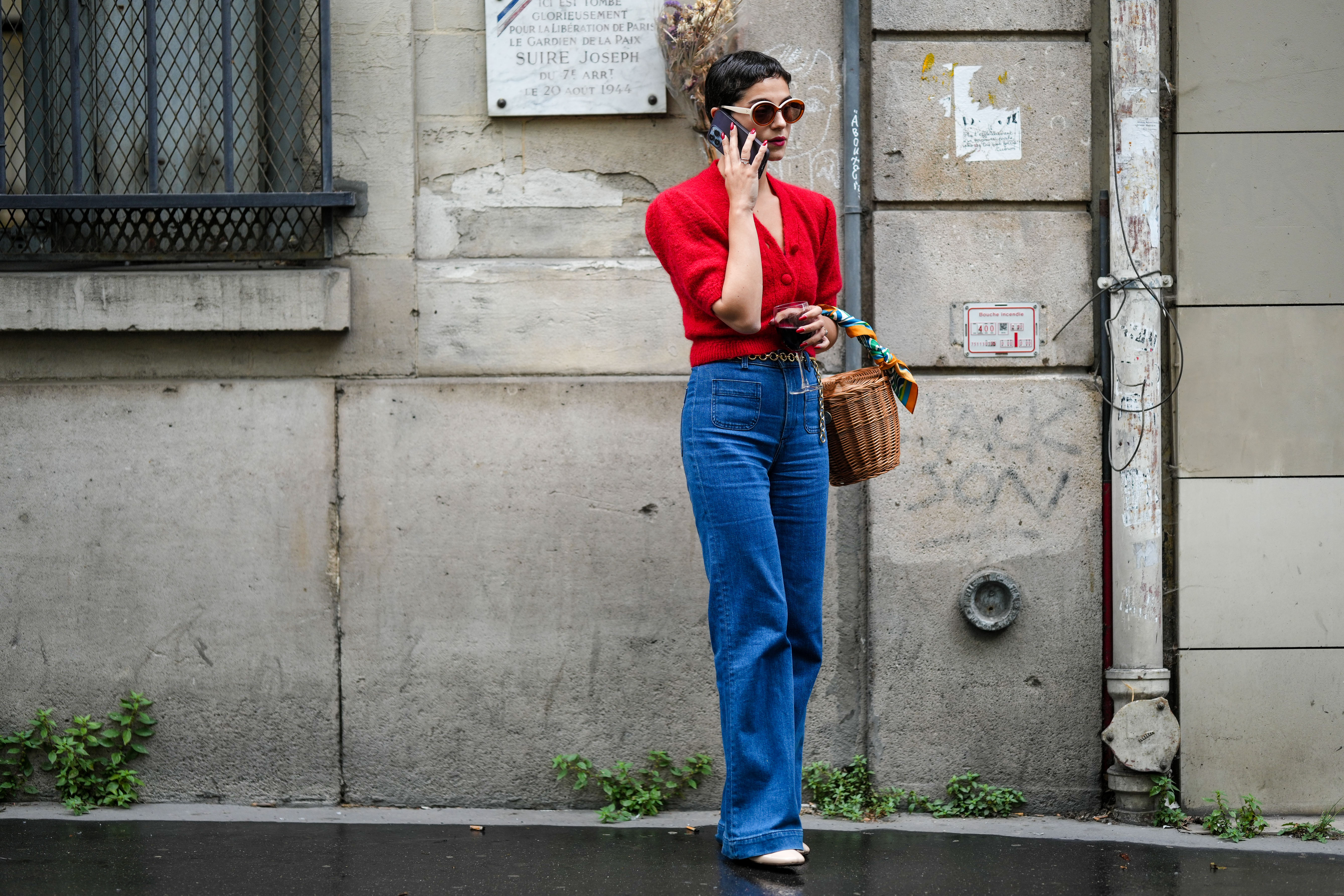 A guest wears circle sunglasses, a red fluffy puffy short sleeves / buttoned cropped cardigan, navy blue denim wide legs pants, a brown wickers basket handbag, a green / blue / orange print pattern silk scarf, rings, white shiny varnished leather heels shoes, a gold chain belt, outside the Hermes show, during Paris Fashion Week - Menswear Spring/Summer 2023, on June 25, 2022 in Paris, France.