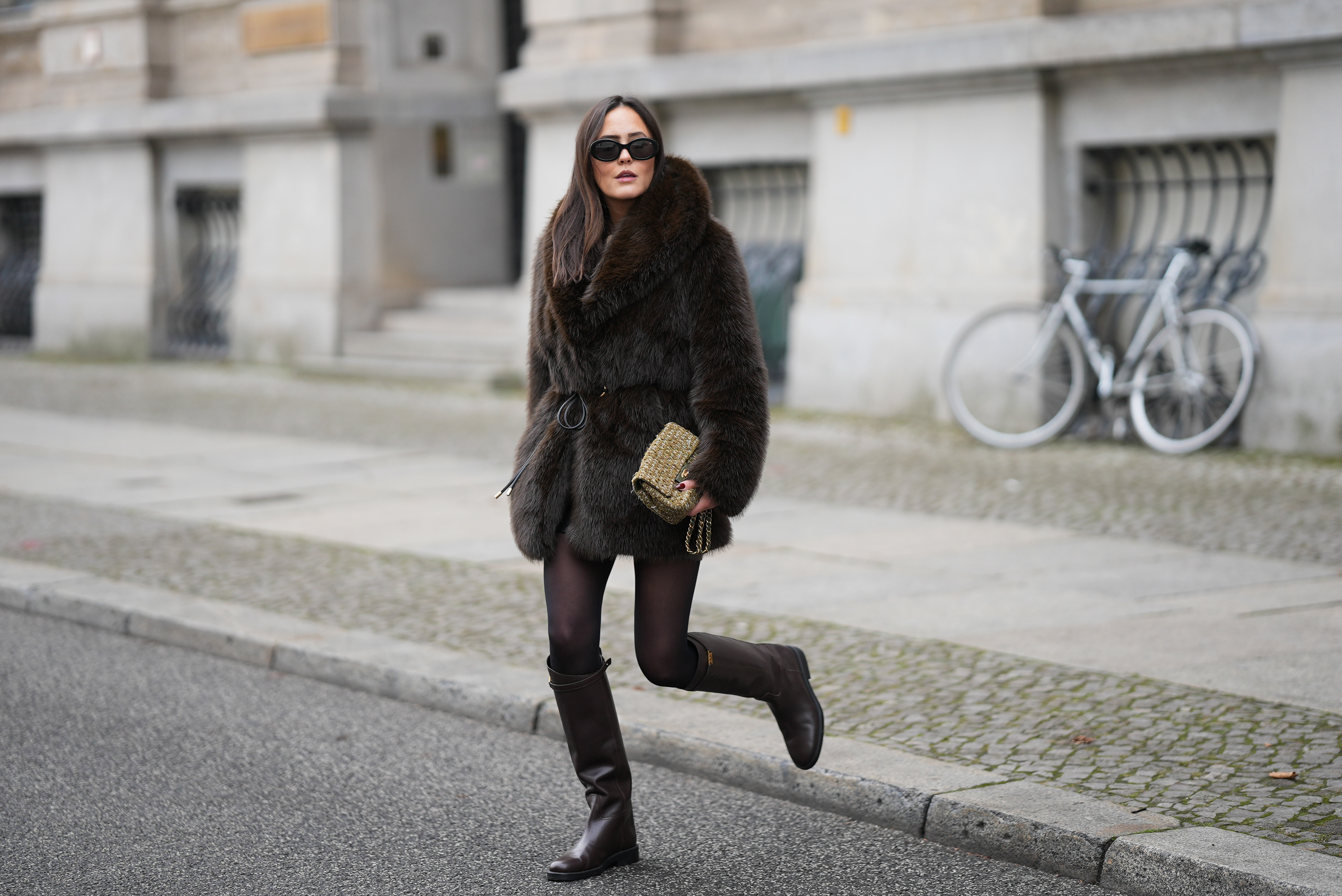 Laura Noltemeyer is seen wearing a dark-brown faux-fur coat with a plush, heavyweight texture, wide notch lapels and a straight hip-length silhouette with a hook and eye belt closing from Zara; a cream knit sweater with a soft ribbed crew neckline from Sézane; semi-sheer black tights; knee-high dark brown leather riding boots with a straight shaft, gold buckle detail and a flat sole from Massimo Dutti; a small quillted and woven flap bag in a khaki green tweed color featuring the signature CC turn-lock and a gold hardware chain strap from Chanel; black oval sunglasses with a glossy acetate frame and dark lenses from Celine; chunky sculptural gold earrings from Zara; her dark brown hair is worn straight with a center part on November 25, 2025 in Berlin, Germany.