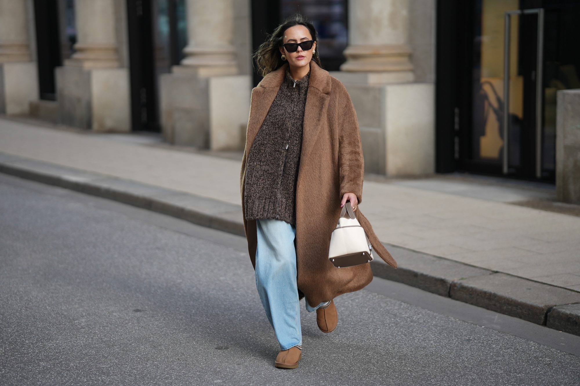 HAMBURG, GERMANY - NOVEMBER 13: Laura Noltemeyer is seen wearing a long camel-colored brushed wool wrap coat with a belt and wide lapels by Max Mara; a chunky zip-front sweater cardigan in a mélange brown wool blend with a textured, hand-knit look by by Aylin Koenig; light blue wide-leg jeans in a soft washed denim with a relaxed fit and front tag detail by Closed; chestnut colored suede slippers with plush lining called „Tasman“ by UGG; a structured bucket-shaped handbag in an off-white linen shade with taupe leather handles and lining details called „Picotin Cargo 18 Toile Goeland“ by Hermès; black cat eye sunglasses with an acetate frame by Yves Saint Laurent; gold statement earrings with a ruffled surface and a green stone element by Mango ; her brown hair is worn loose in soft waves with a center part on November 13, 2025 in Hamburg, Germany. (Photo by Moritz Scholz/Getty Images)