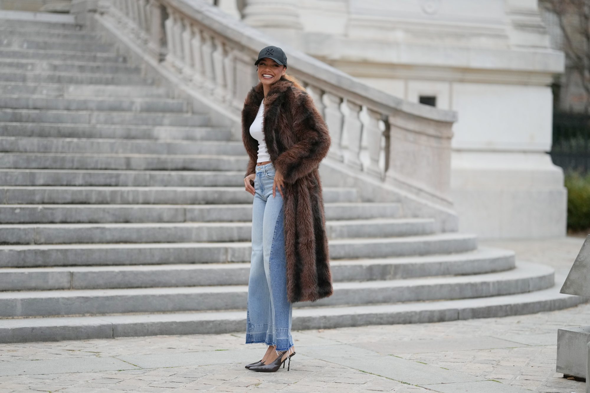 PARIS, FRANCE - DECEMBER 11: Alicia Aylies wears hair pulled back into a sleek low ponytail, natural skin with defined brows and lipstick, a black baseball NY cap, a white fitted cropped top by Celine, a long brown faux fur coat with a straight cut and button closure, light blue high waist flared wide leg jeans with a raw hem, and dark pointed toe high heel pumps shoes, during a street style fashion photo session, on December 11, 2025 in Paris, France. (Photo by Edward Berthelot/Getty Images)