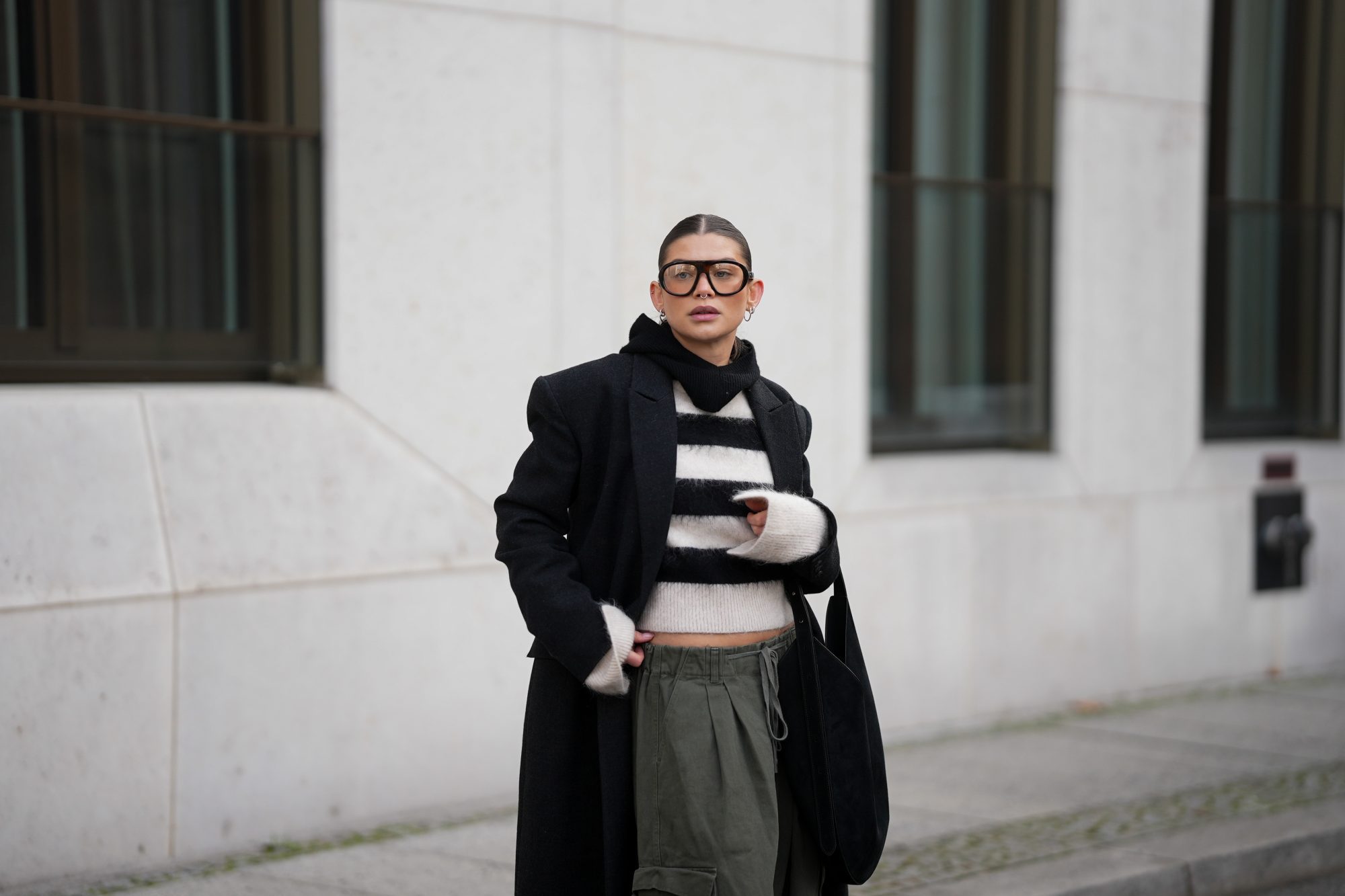 BERLIN, GERMANY - NOVEMBER 25: Sophia Geiss is seen wearing an oversized long black wool coat with structured shoulders and a straight, ankle-length silhouette from Herskind; a slightly cropped, fuzzy black-and-white striped knit sweater from H&M; wide-leg khaki green cargo trousers with drawstring waist and large side pockets from The Frankie Shop; a black suede crescent shoulder hobo bag with a minimalist curved shape from Aesther Ekme; oversized aviator glasses with tortoiseshell frame from Prada; a black knit balaclava from COS; small silver hoop earrings from Maria Black; her brown hair is styled in a sleek low bun with a center parting on November 25, 2025 in Berlin, Germany. (Photo by Moritz Scholz/Getty Images)