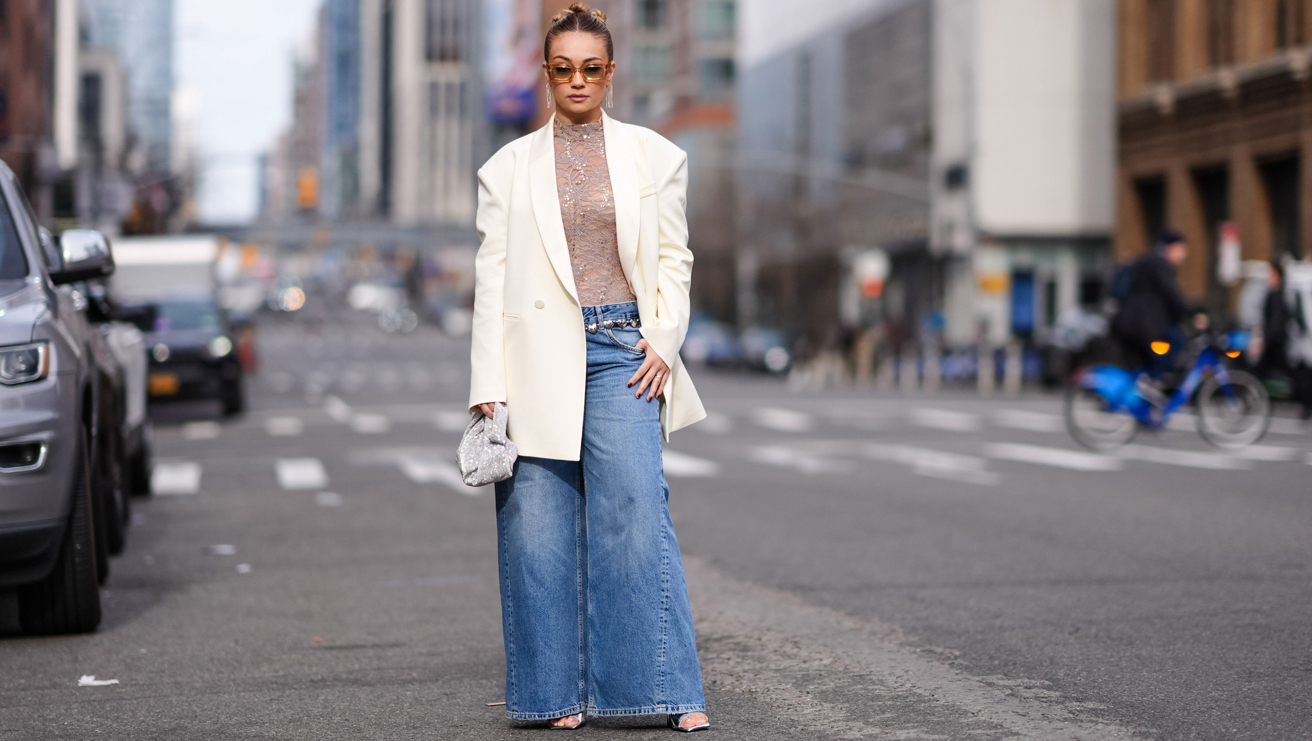 NEW YORK, NEW YORK - FEBRUARY 11: Merve Gorgotz wears sunglasses, earrings, a floral embroidery gray turtleneck top, a white oversized blazer jacket flared blue denim jeans pants , silver shoes, outside Area, during New York Fashion Week, on February 11, 2024 in New York City. (Photo by Edward Berthelot/Getty Images)