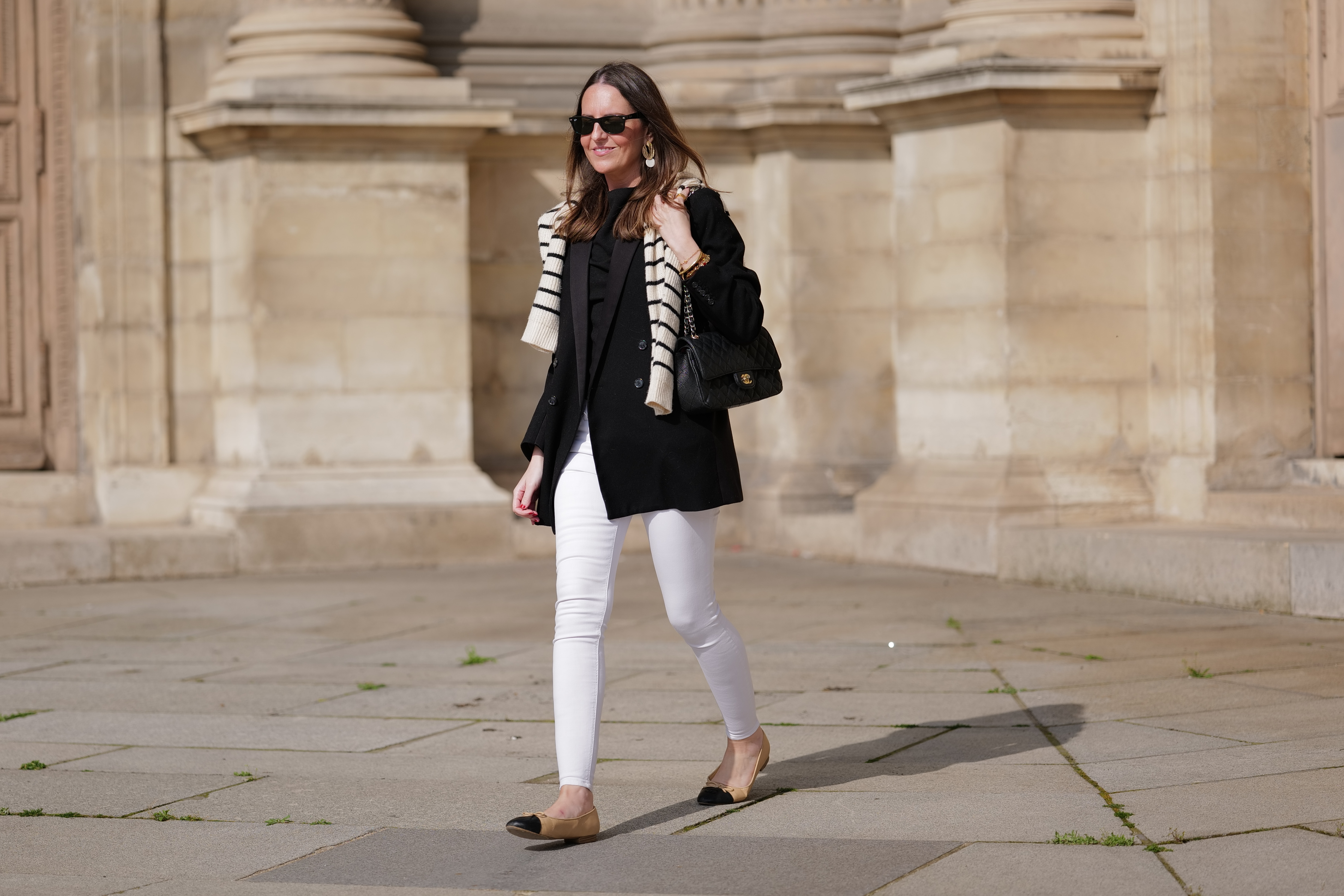Alba Garavito Torre wears black sunglasses from Ray Ban, gold and white pendant large earrings, a black t-shirt, a white with black small striped print pattern wool pullover from Massimo Dutti, a black shiny grained leather Timeless shoulder bag from Chanel, a black oversized blazer jacket from Munthe, white skinny denim pants from Zara, beige shiny leather with black toe-cap ballerinas from Chanel , during a street style fashion photo session, on April 29, 2023 in Paris, France.