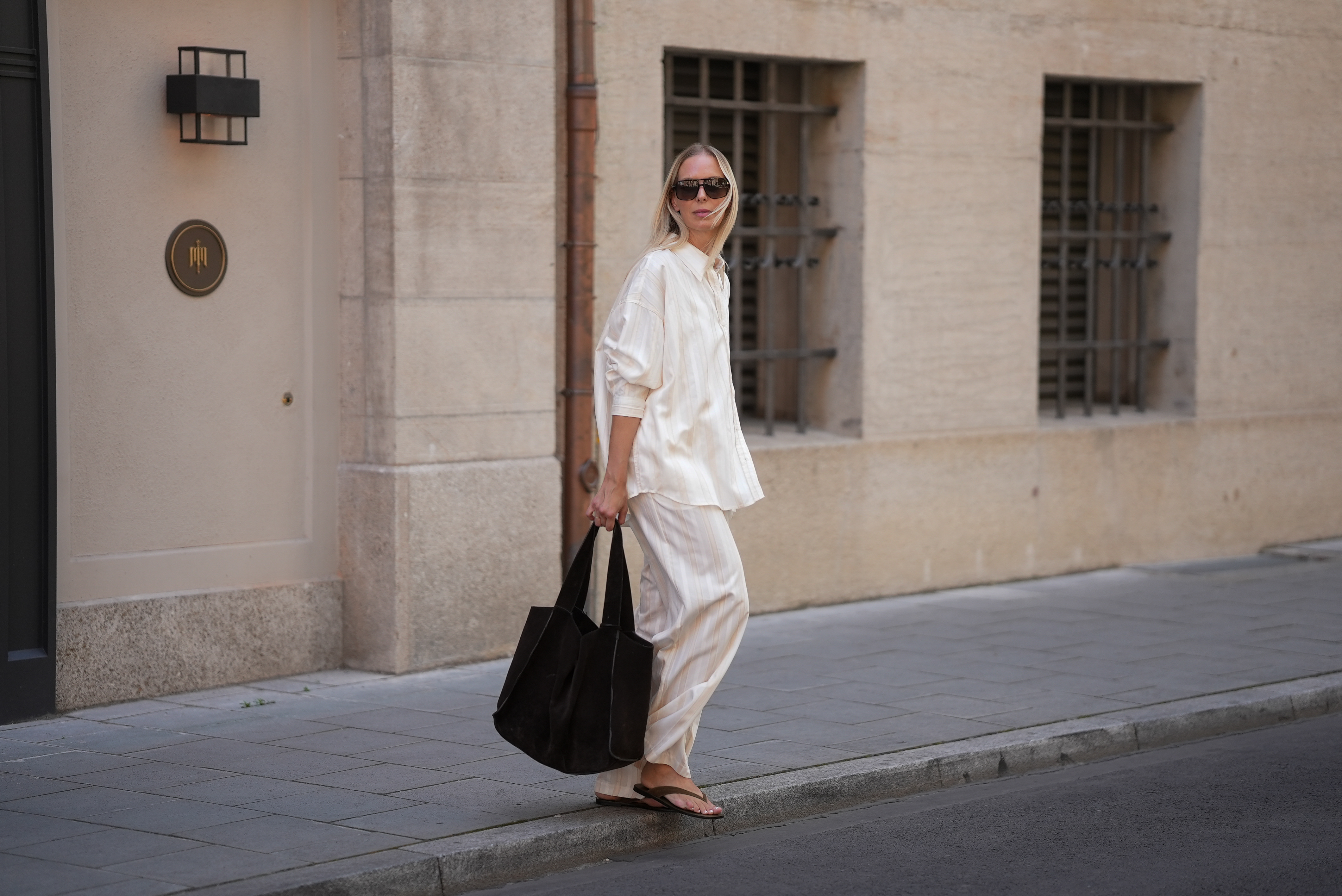 Mirja Klein is seen wearing a matching set consisting of a white striped oversized dobby-weave shirt with collar and buttons down in the front from H&M and a white striped straight pull-on trousers with loose fit and straight legs from H&M; a black suede tote bag with slouchy design and open top from COS; black oversized aviator sunglasses from Tom Ford; a pair of khaki green suede flip flops from A.EMERY on June 18, 2025 in Munich, Germany.