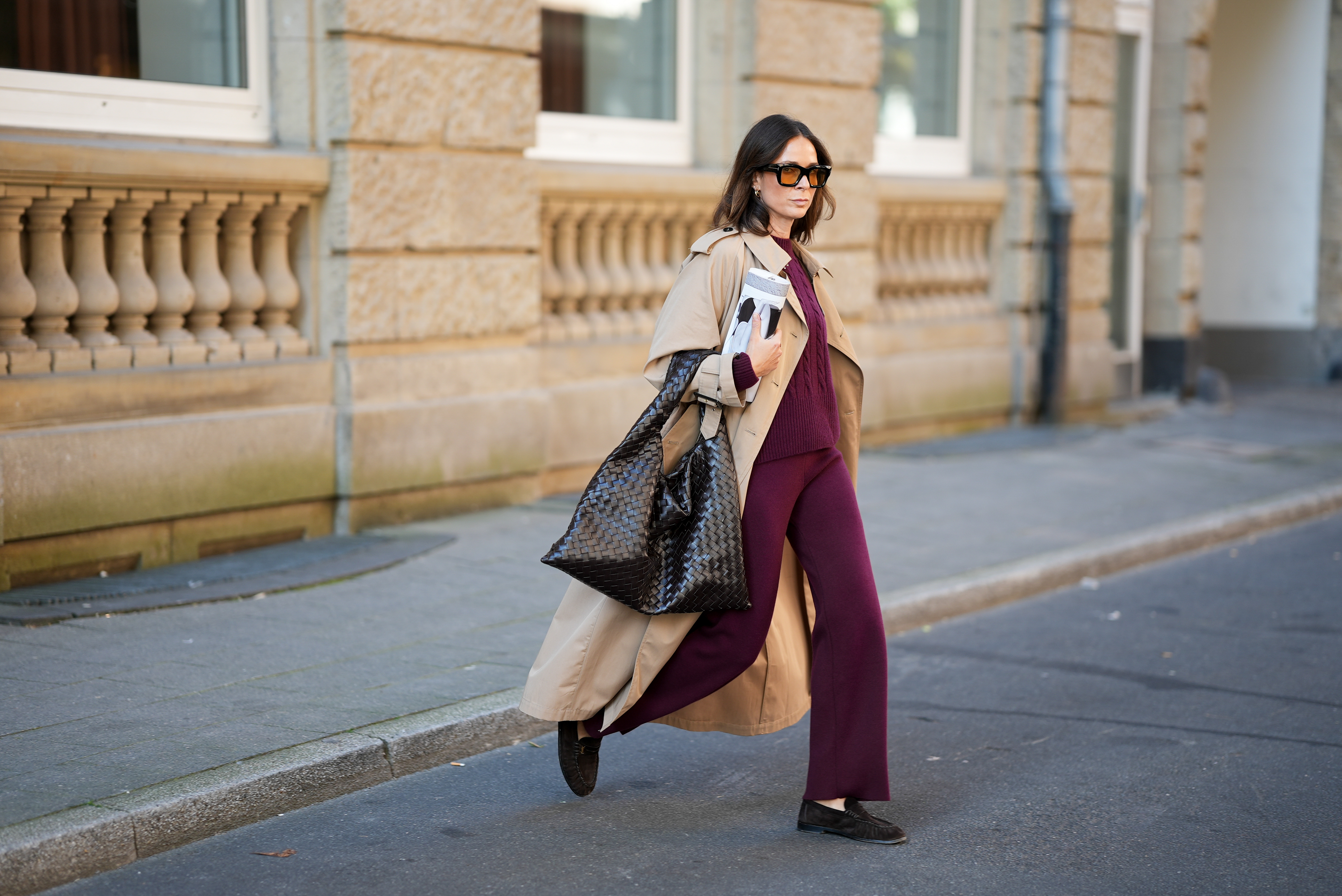 Patricia Wirschke is seen wearing a classic beige long Herno trench coat, with a notched collar, storm flap, and belted cuffs; a burgundy Incentive co ord set with a cable-knit sweater, featuring a ribbed hem, cuffs, and neckline and wide-leg knit pants with a relaxed fit; a pair of Le Loafer soft suede Saint Laurent moccasins in dark brown with golden logo detail on the side; a dark brown Hop bag from Bottega Veneta with the famous Intrecciato leather construction, woven; Tom Ford Ilias squared frame black sunglasses with orange toned lens; Bvlgari Serpenti Viper golden earrings; her brown hair is styled in a straight, shoulder-length look with a middle part; she holds a rolled up magazine in her hand on August 19, 2025 in Duesseldorf, Germany.