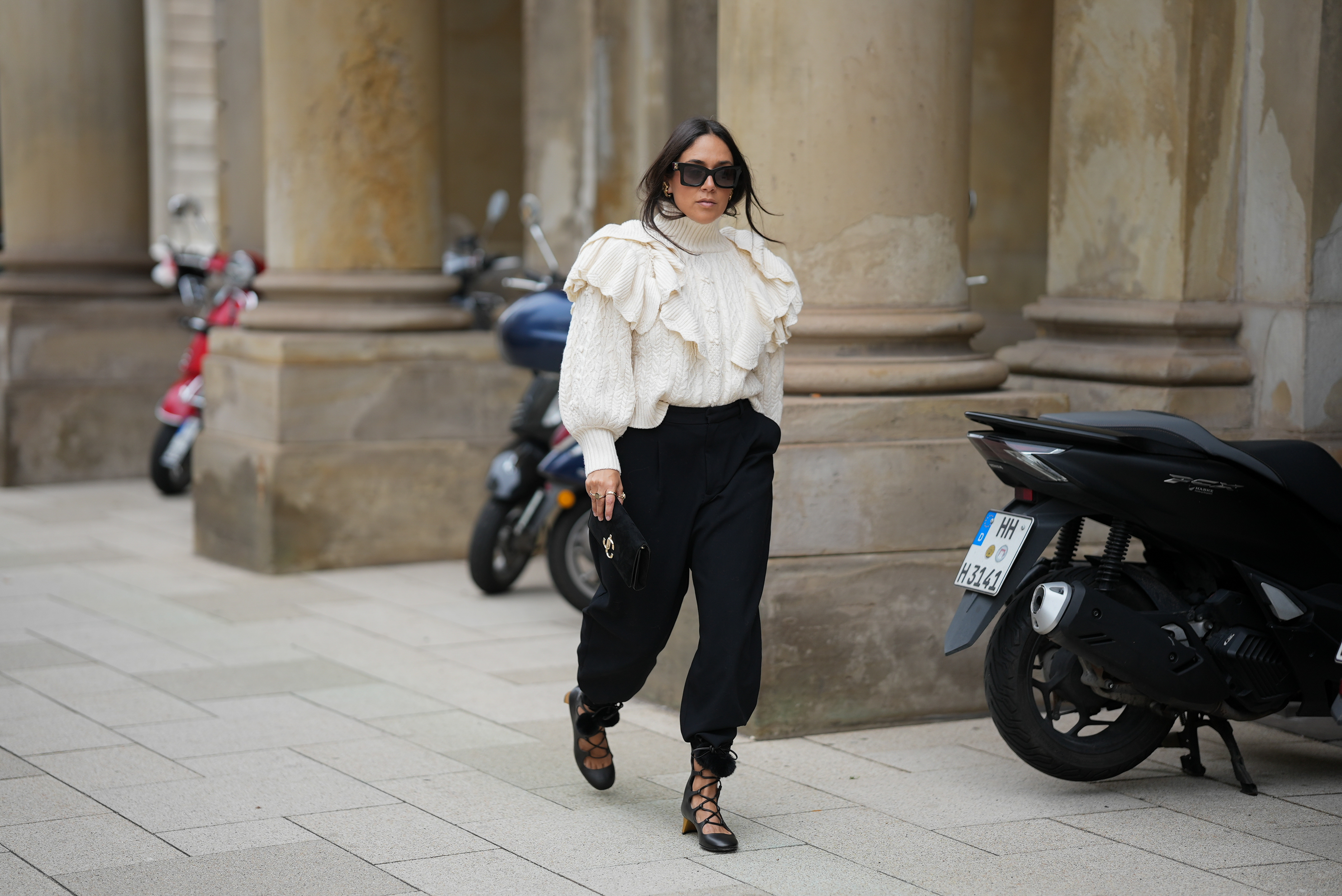 Jennifer Casimiro is seen wearing a cream cable-knit turtleneck sweater with voluminous ruffled detailing on the shoulders from Zara; black high-waisted trousers with a tapered silhouette, cinched at the ankles from Zara; black lace-up shoes with block heels, featuring pom-pom accents at the ankle ties from Gucci; the Suede Varenne small black clutch bag with a rectangular shape and embossed gold branding from Jimmy Choo; oversized black rectangular sunglasses from MiuMiu; gold cuffed earrings from Zara; her black hair is worn down with a center part on September 19, 2025 in Hamburg, Germany.