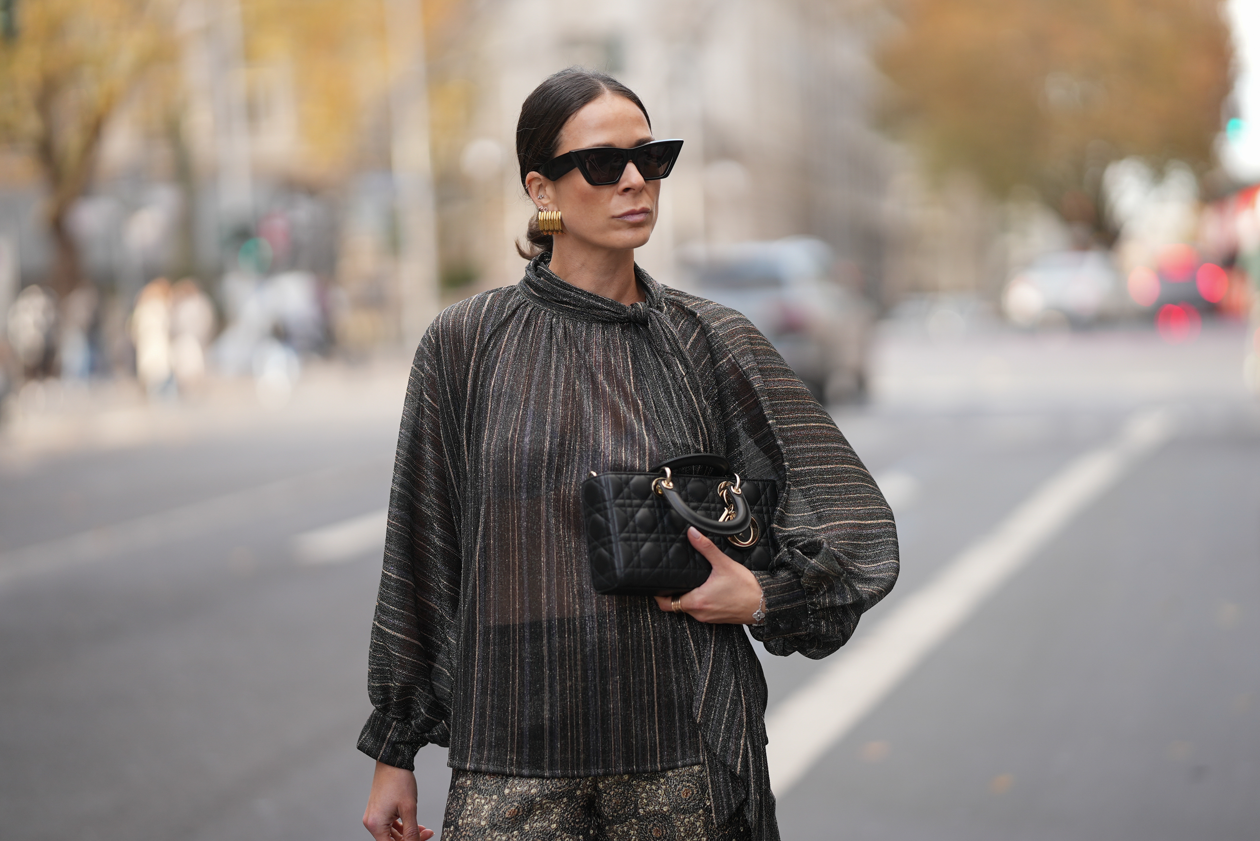 Patricia Wirschke is seen wearing a shimmering metallic striped blouse in shades of charcoal, bronze, and silver with a see through, flowing silhouette, high neckline and tie detail at the collar by Si Yu; paired with satin wide-leg trousers featuring an intricate geometric print in gold and dark brown tones by Si Yu; a small black quilted leather “Lady D-Joy” handbag with gold hardware by Dior; black cat-eye sunglasses with a chunky rim by Phoebe Philo; gold statement hoop earrings by an unknown brand; a rose gold “B.zero1” ring by Bulgari; a white gold “Alhambra” bracelet by Van Cleef & Arpels; her brown hair is in a sleek low bun parted in the middle on November 10, 2025 in Duesseldorf, Germany.