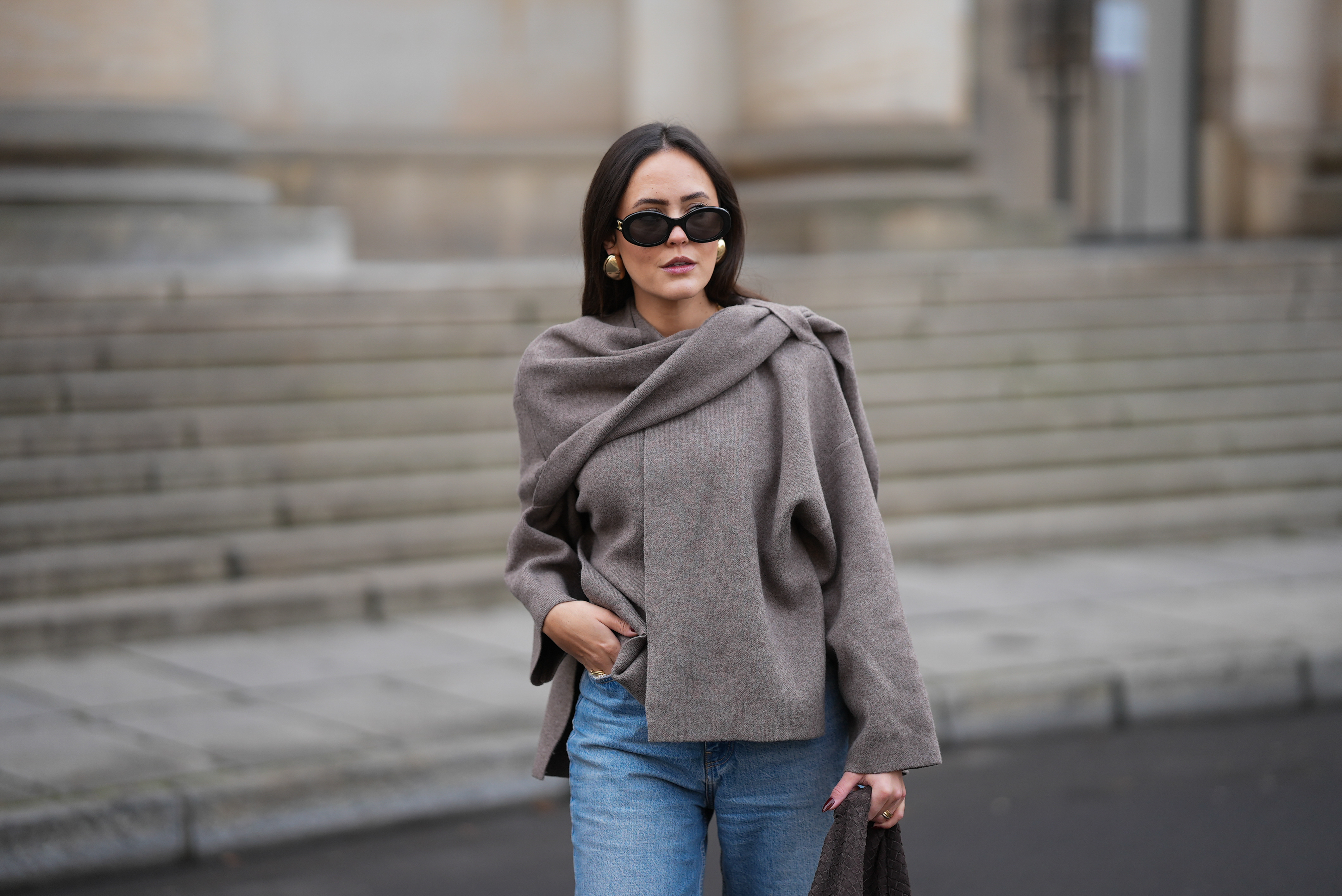 Laura Noltemeyer is seen wearing a taupe fine-knit sweater with a sculptural draped front collar from Zara; light-blue straight-leg denim jeans with a soft fading from Zara; beige suede winter boots with a rounded toe, rope laces, tonal stitching and a thick rubber sole from Inuikii;a large textured dark-brown shoulder bag in a soft sculptural, slouchy shape and woven leather from Les Visionnaires x Michi from the Want; black oval sunglasses with a glossy acetate frame and dark lenses from Celine; chunky gold sculptural earrings from Zara; dark brown soft leather gloves from Zara; her dark brown hair is worn straight with a middle parting, tucked behind the ears on November 25, 2025 in Berlin, Germany.