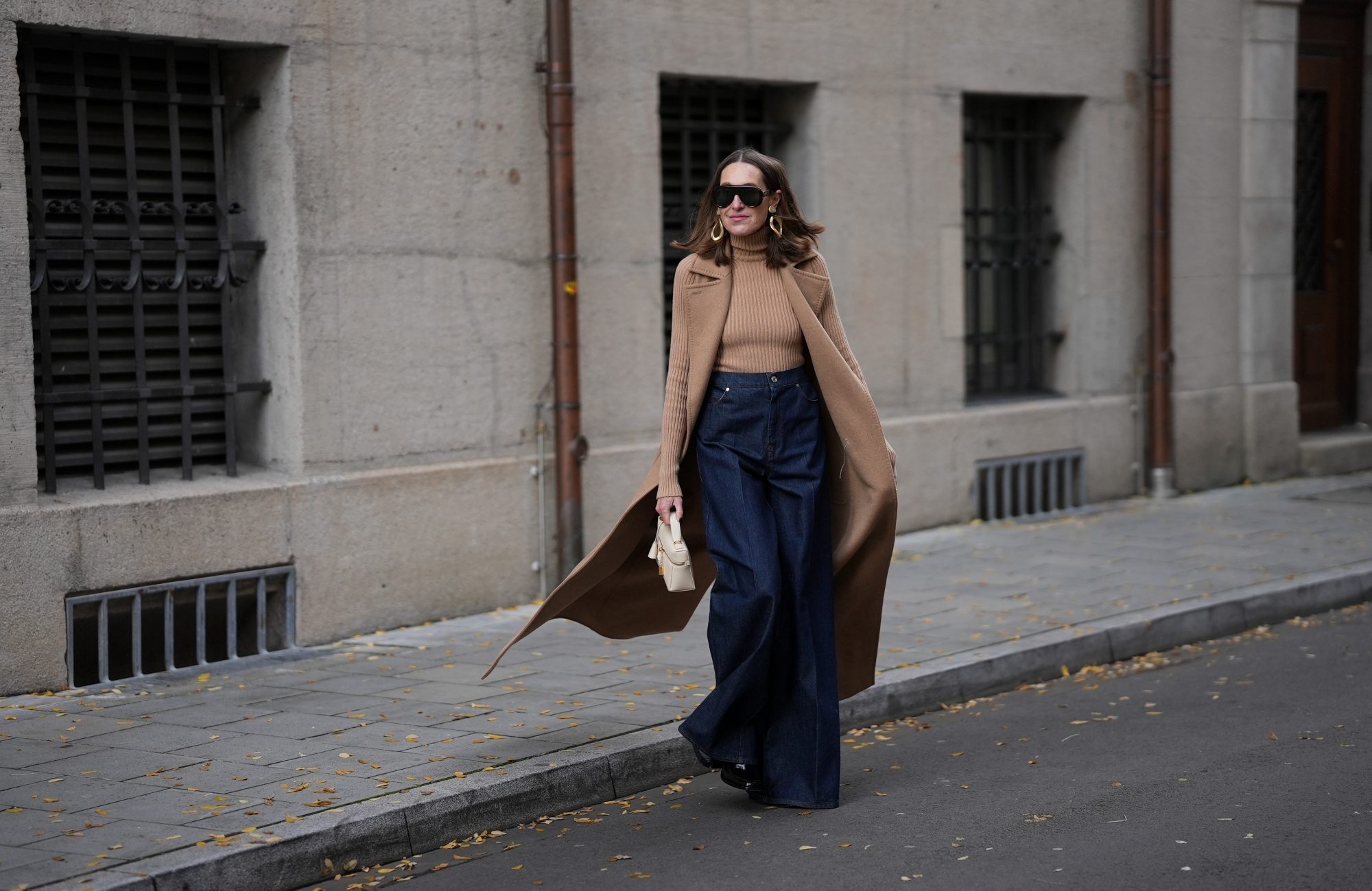 MUNICH, GERMANY - OCTOBER 28: Annette Weber is seen wearing a long camel sleeveless wool coat with wide notch lapels, oversized patch pockets and a fluid silhouette from Max Mara; a fitted camel rib-knit turtleneck body with a fine vertical texture from Max Mara; dark blue wide leg denim pants with a high waist and a structured drape from Max Mara; black patent boots with a shiny finish and chunky heel from Jimmy Choo; a small cream structured leather bag featuring a small top handle, gold zip closure and a minimal trapeze silhouette from Loro Piana; oversized black sunglasses with a bold shield frame and dark lenses from Gucci; large sculptural gold-tone statement earrings with organic round shapes from Max Mara; her brown hair is worn straight and middle parted on October 28, 2025 in Munich, Germany. (Photo by Moritz Scholz/Getty Images)