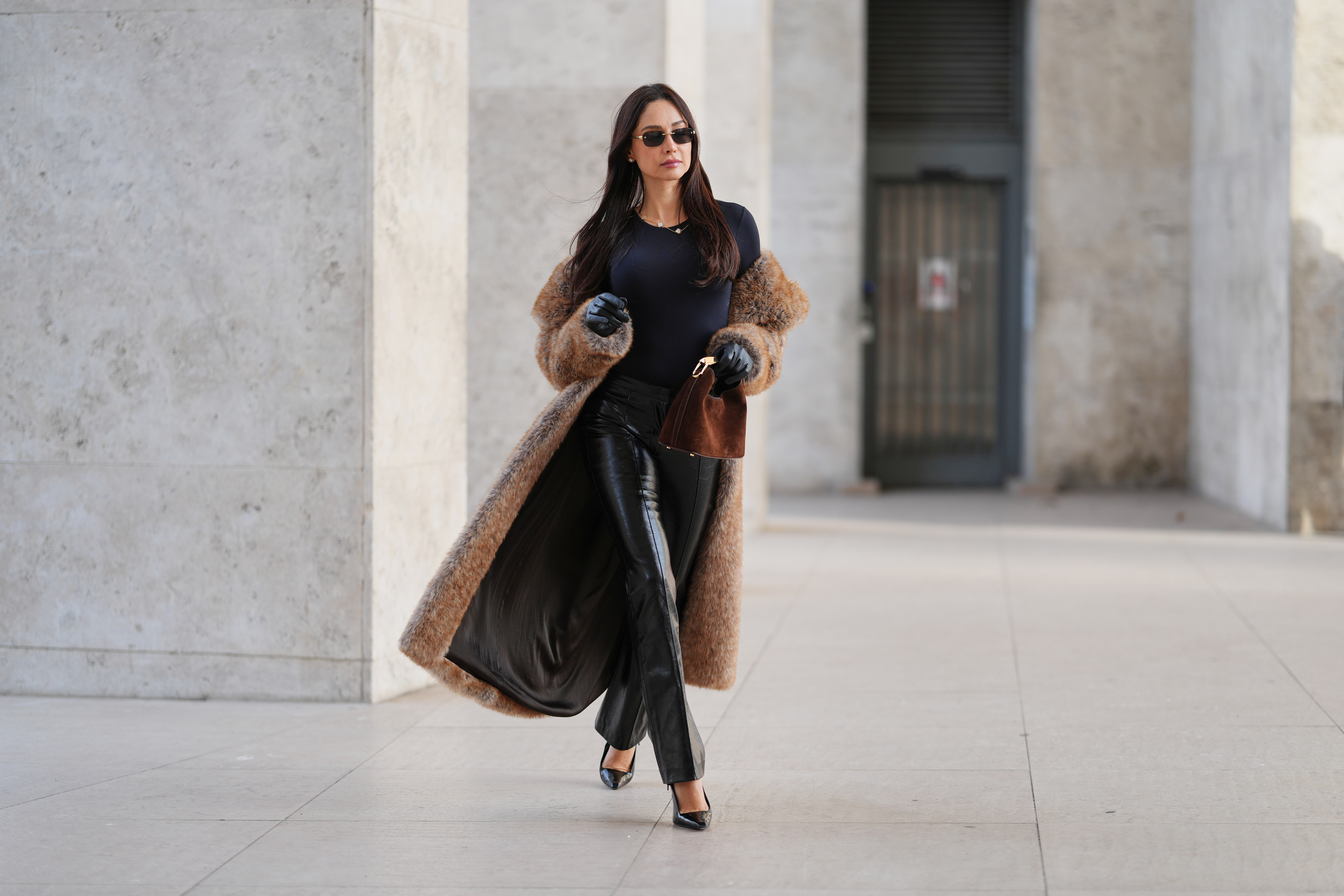 Patricia Gloria Contreras wears long straight dark brown hair worn loose, natural skin with neutral lipstick, narrow rectangular black sunglasses, a long brown faux fur coat by The Frankie Shop worn open, a black fitted long sleeve top with a round neckline, black high waisted straight leg leather pants by Mango, black leather gloves, black pointed toe high heel pumps by Saint Laurent, and a brown suede mini handbag with a curved top handle by Elleme, during a street style fashion photo session, on December 09, 2025 in Paris, France.