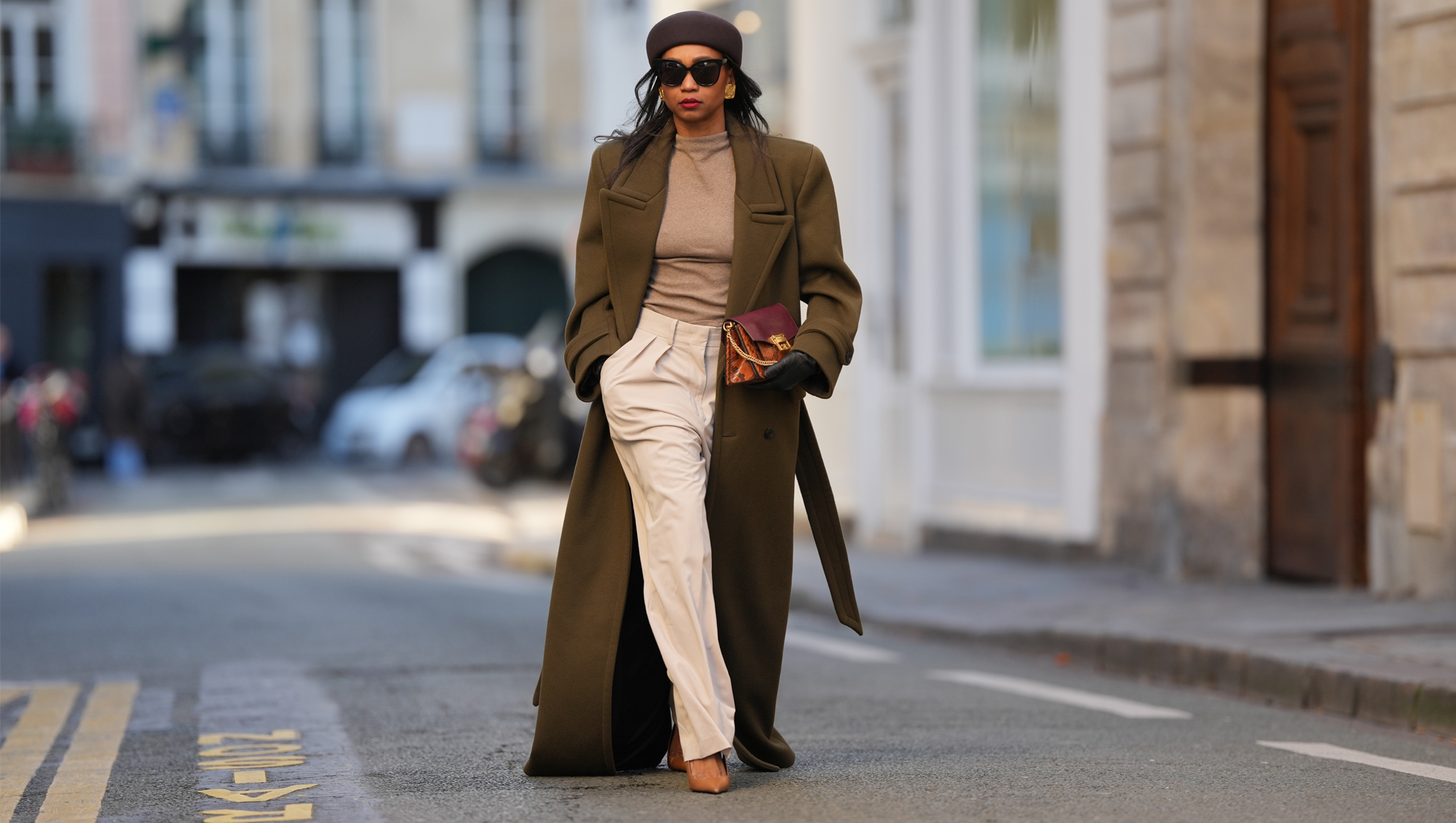PARIS, FRANCE - JANUARY 03: Emilie Joseph wears straight black hair worn loose under a brown wool pillbox hat by COS, black rectangular sunglasses, a natural makeup look with defined brows, even skin, deep red lipstick, gold-tone statement earrings, an oversized military-style wool long coat in deep olive green with sharp structured shoulders by Saint Laurent, a fitted taupe mock-neck knit top, high-waisted wide-leg cream white canvas trousers with front pleats by The Frankie Shop, a burgundy and python-embossed leather clutch bag with a gold-tone buckle by Givenchy, black leather gloves, pointed-toe leather slingback heels shoes in cognac brown by Saint Laurent, during a street style photo session, on January 03, 2026 in Paris, France.