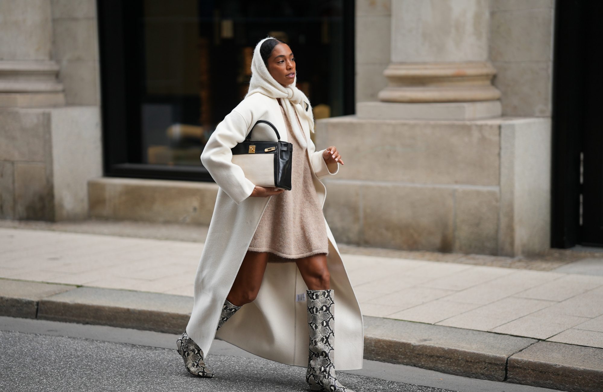 HAMBURG, GERMANY - OCTOBER 16: Cherifa Akili is seen wearing an oversized cream wool coat with a clean straight cut and dropped shoulders from Anine Bing; a wide beige knit sweater mini dress with a soft and fuzzy cashmere texture from by Aylin Koenig; cream knit headscarf in a fluffy mohair-blend from by Aylin Koenig; knee-high snake-embossed leather boots in a grey and black reptile pattern with a pointed toe from Arket; a structured two tone &bdquo;Kelly bag&ldquo; with a structured black leather body, cream linen canvas front detailing and gold hardware from Herm&eacute;s; her hair is styled slicked back with middle parting and partly covered by the hood-scarf on October 16, 2025 in Hamburg, Germany. (Photo by Moritz Scholz/Getty Images)