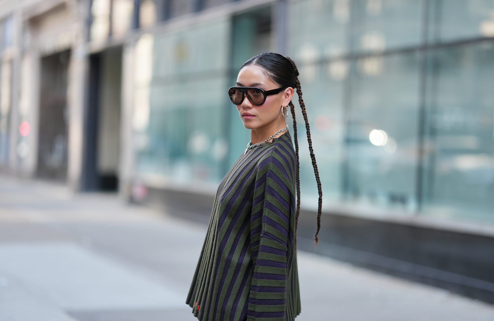 NEW YORK, NEW YORK - SEPTEMBER 14: A guest wears a center-parted slick low bun, oversized dark brown gradient sunglasses, a chunky silver chain necklace with a small round silver pendant and medium silver hoop earrings, a ribbed knit long-sleeve sweater with vertical dark navy and olive green stripes over olive green shorts, outside Gabe Gordon, during New York Fashion Week, on September 14, 2025 in New York, New York (Photo by Edward Berthelot/Getty Images)