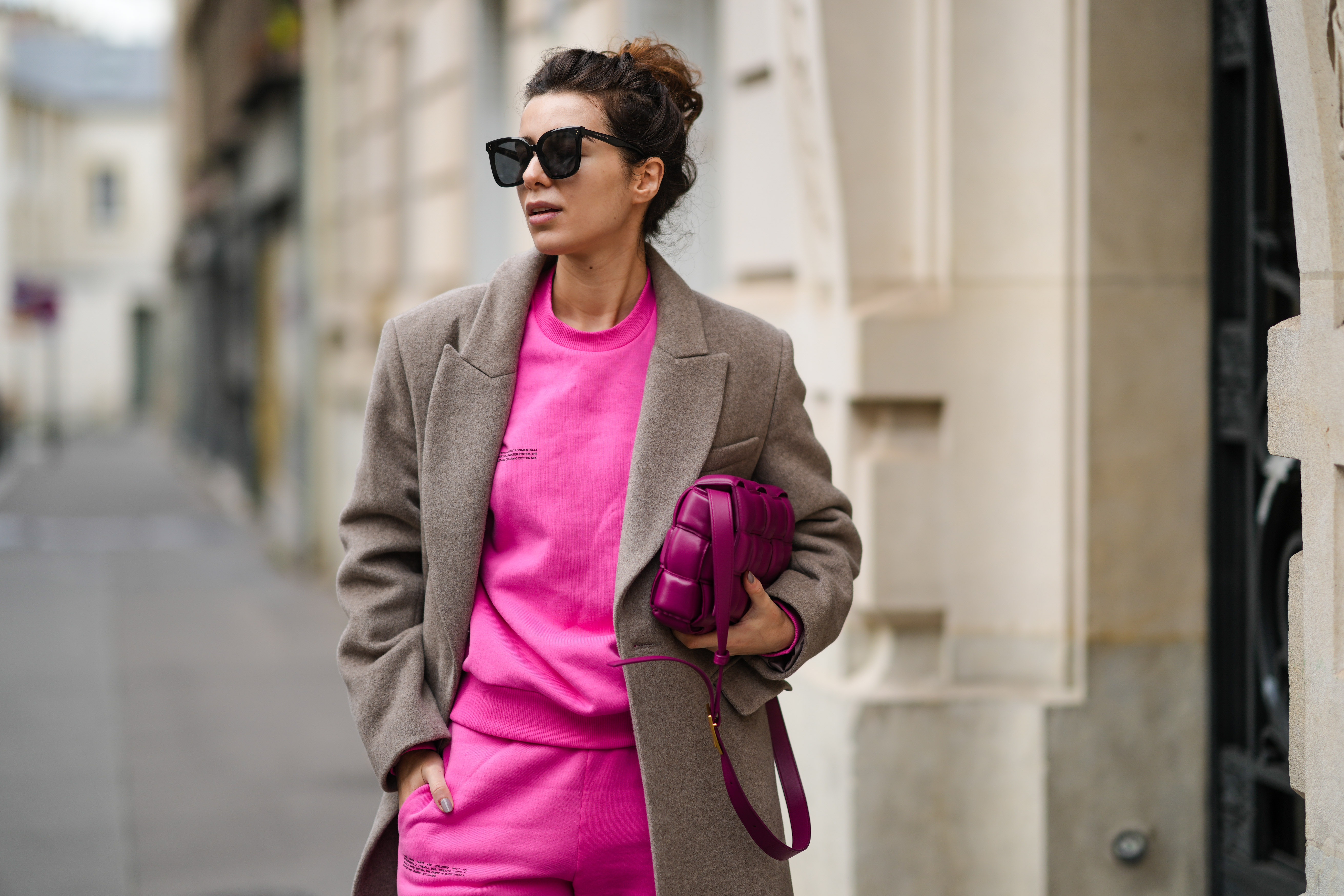 Katie Giorgadze @katie.one wears a black sunglasses, a neon pink recycled cotton sweater, matching neon pink recycled cotton sport pants / jogger pants, a pale brown wool long coat, a purple shiny leather Padded Cassette shoulder bag from Bottega Veneta, during a street style fashion photo session, on December 11, 2021 in Paris, France.