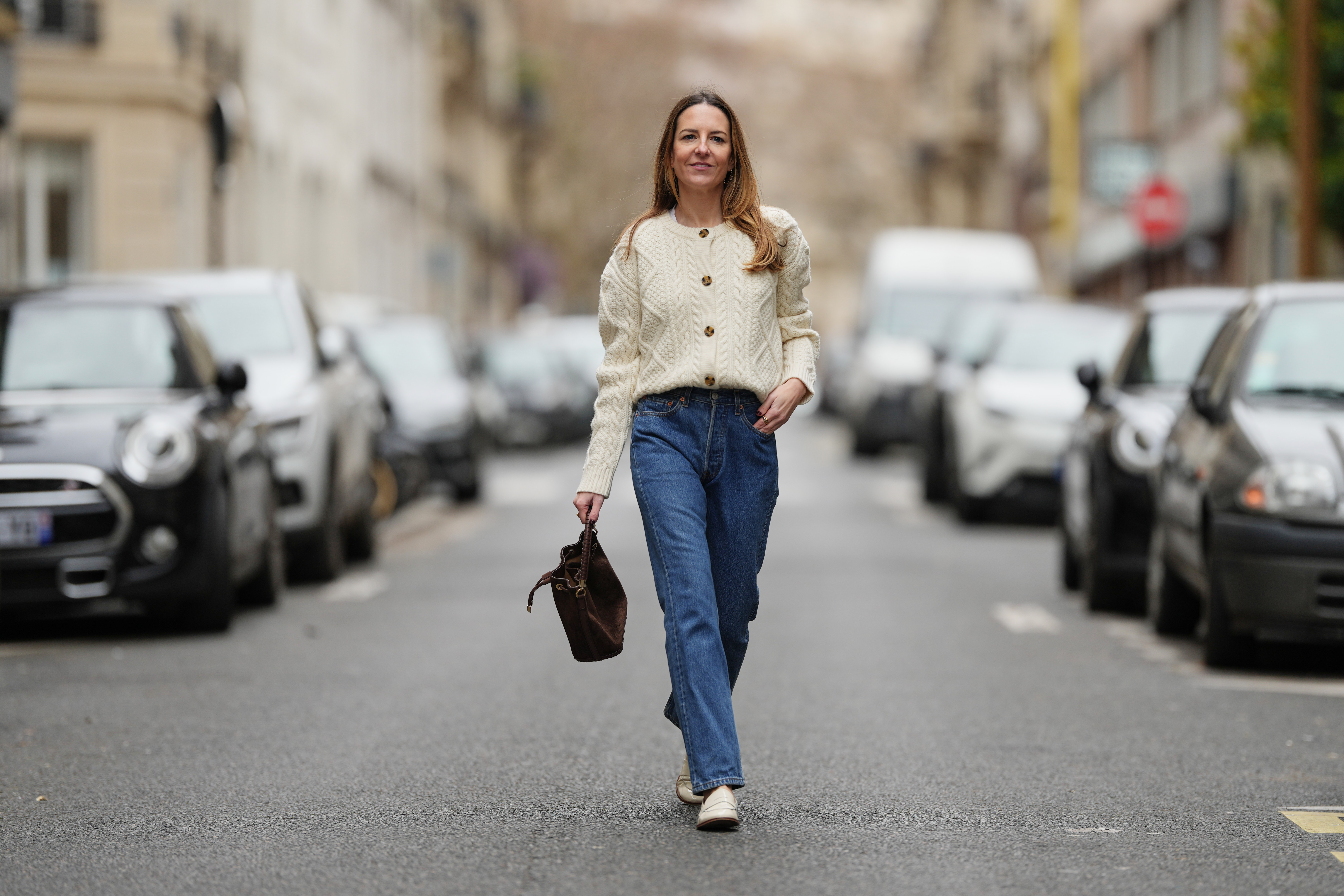 Alba Garavito Torre wears long light brown hair worn loose and parted at the side, small gold-tone earrings by Sempiterno Paris, an ecru white woven cable-knit cardigan with a round neckline, textured panels and front button fastening, dark blue straight-leg denim 501 jeans by Levis with a classic five-pocket construction, ecru patent leather loafers by Jonak with a rounded toe and low heel, a chocolate brown suede bucket bag by Saonara with a drawstring closure and top handle carried by hand, during a street style fashion photo session, on January 15, 2026 in Paris, France.
