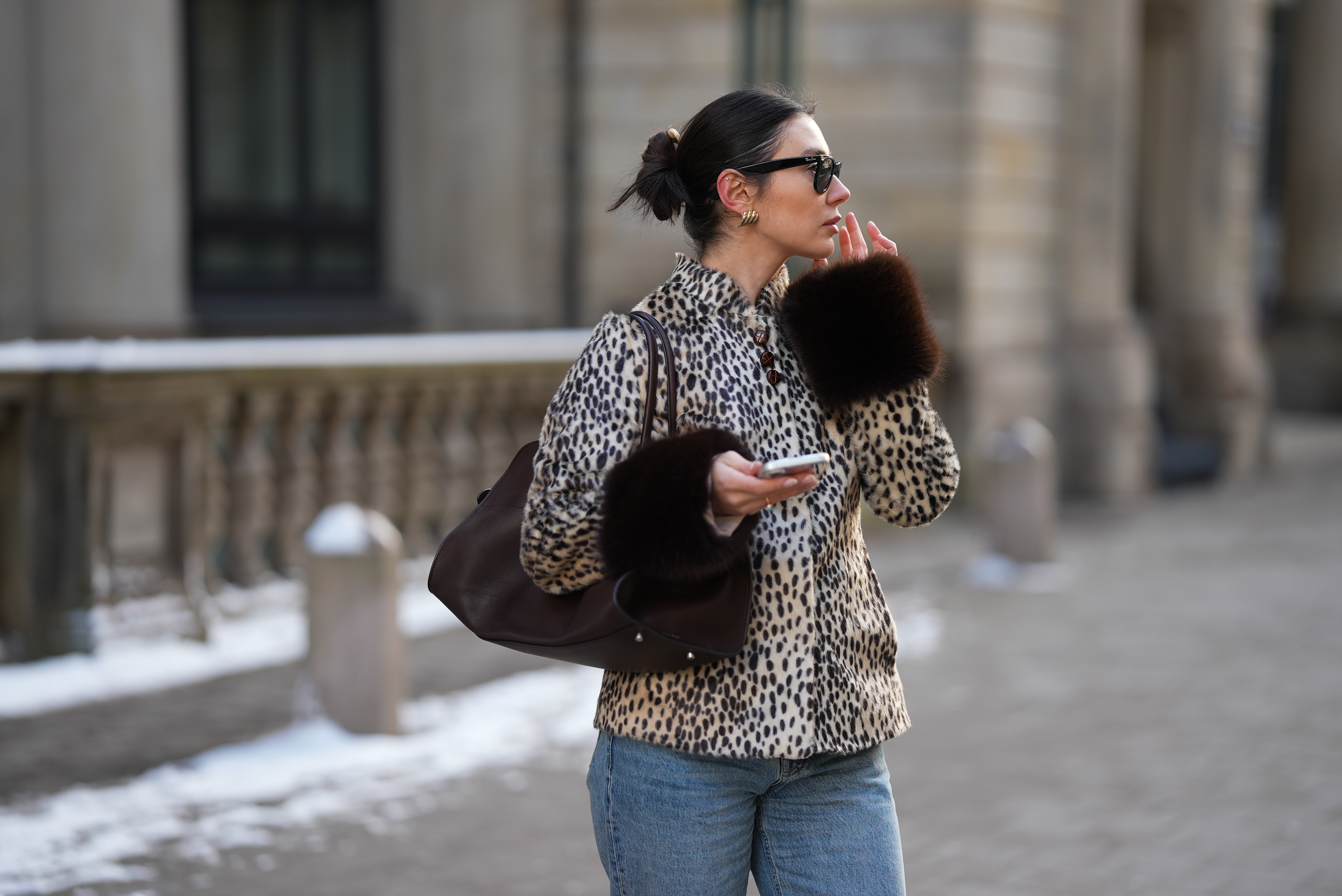Milena Karl is seen wearing a cream and black leopard print "dolly" faux fur jacket with a classic collar, front leather button closure and dark brown fur trimmed cuffs from Carelli; light blue straight leg jeans with a mid rise fit from Massimo Dutti; the &ldquo;Soft Margaux 15&rdquo; handbag crafted in smooth dark brown leather with a softly structured silhouette, side buckle detailing, curved top handles and subtle logo detailing from The Row; holding an iPhone from Apple; rectangular black "Wayfarer" sunglasses with dark tinted lenses from Ray-Ban; sculptural gold colored earrings from Mango; her dark brown hair styled in a sleek low bun on February 14, 2026 in Hamburg, Germany.