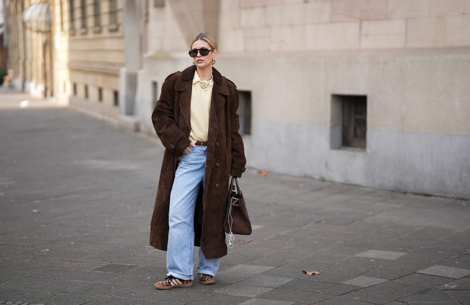 FRANKFURT AM MAIN, GERMANY - JANUARY 21: Julia Kammerer is seen wearing a long chocolate brown suede vintage trench coat with a wide lapel collar, button front and a relaxed fit; a butter yellow blouse with classic collar from JJXX; a light yellow knit sweater from H&M layered above the blouse; light blue wash jeans with turned up cuffs, straight leg and relaxed fit from Levi’s; a mocha brown suede leather Midi Hudson tote bag with top handle and adjustable belt from DeMellier; oversized aviator sunglasses with brown tortoiseshell frames and tinted lenses from H&M; a gold-plated brass necklace with natural freshwater pearls from Scandivv; a golden chain necklace with key lock pendant from MyJewellery; a golden chain necklace with „A“-letter pendant from Scandivv; chunky golden hoop earrings from COS; a slim brown leather belt with golden buckle from Lilysilk; Samba OG leather-trimmed leopard-print pony hair sneakers from Adidas x Wales Bonner; her blonde hair styled back on January 21, 2026 in Frankfurt am Main, Germany. (Photo by Moritz Scholz/Getty Images)
