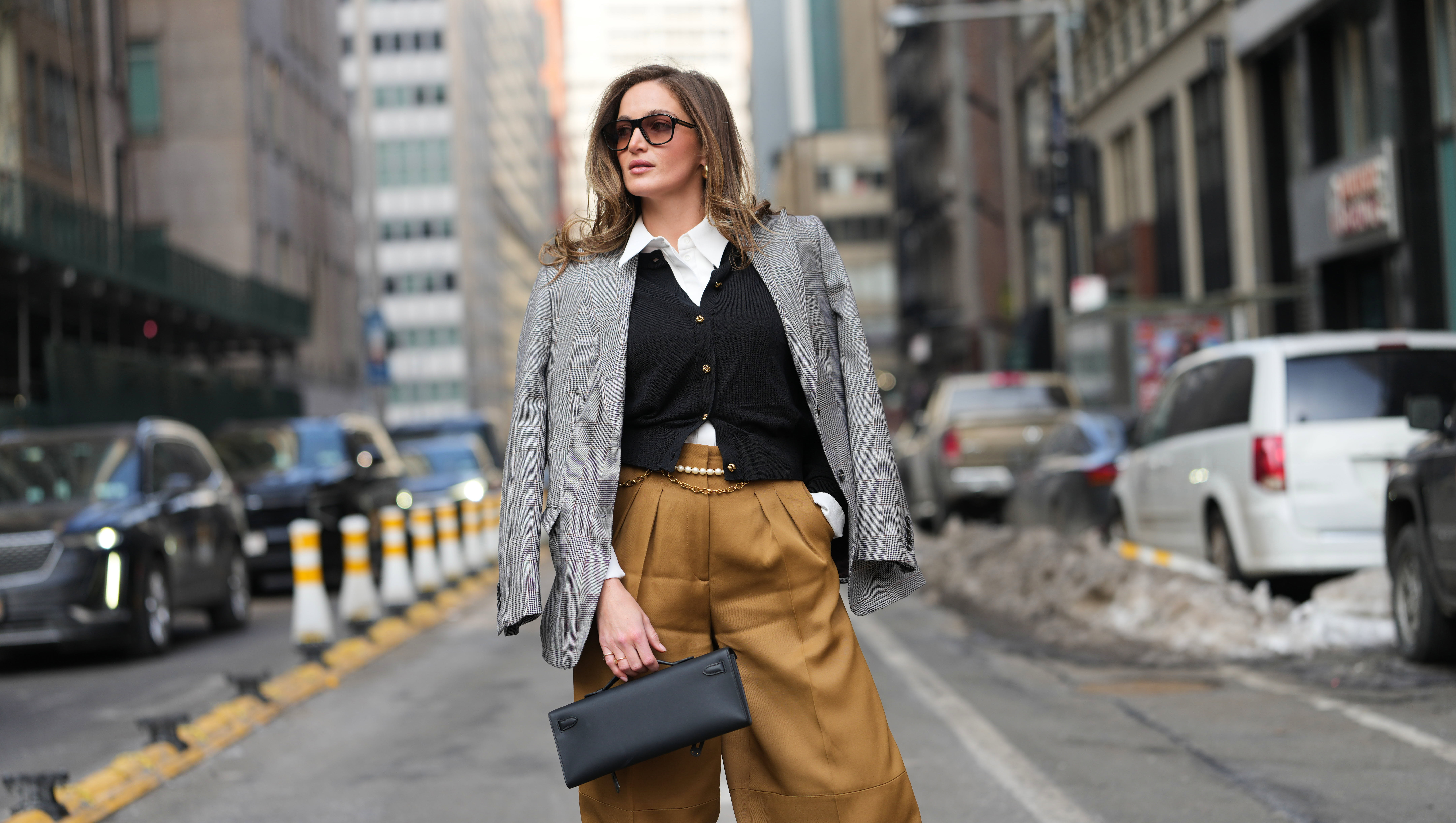 NEW YORK, NEW YORK - FEBRUARY 14: A guest wears loose hair, black sunglasses, gold earrings, rings, a black leather Kelly clutch bag from Hermes, a gray plaid blazer jacket, a black knit V-neck cardigan with gold buttons, a white collared shirt, brown wide-leg pants, a gold chain belt, a white pearls belt, outside Altuzarra, during New York Fashion Week, on February 14, 2026 in New York City (Photo by Edward Berthelot/Getty Images)