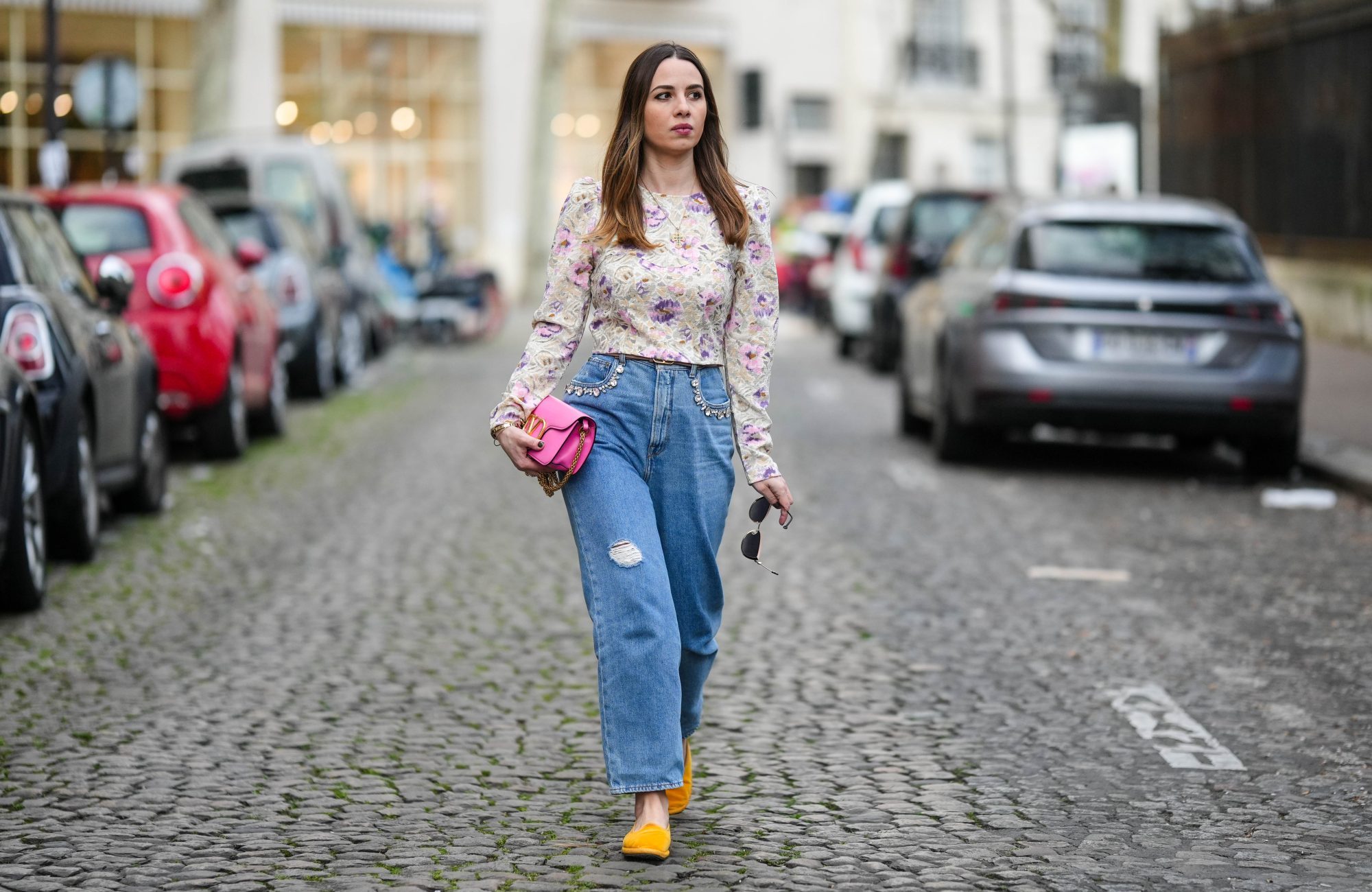 PARIS, FRANCE - MARCH 25: Maria Rosaria Rizzo wears a gold long chain with large cross pendant necklace, a beige with purple and pink tie and dye print pattern long sleeves / puffy shoulder blouse, a neon pink shiny leather long clutch with gold large logo buckle from Valentino, a brown beige shiny leather with gold mini logo buckle belt from Valentino, blue faded denim ripper large pants, yellow gold velvet loafers , during a street style fashion photo session, on March 25, 2023 in Paris, France. (Photo by Edward Berthelot/Getty Images)