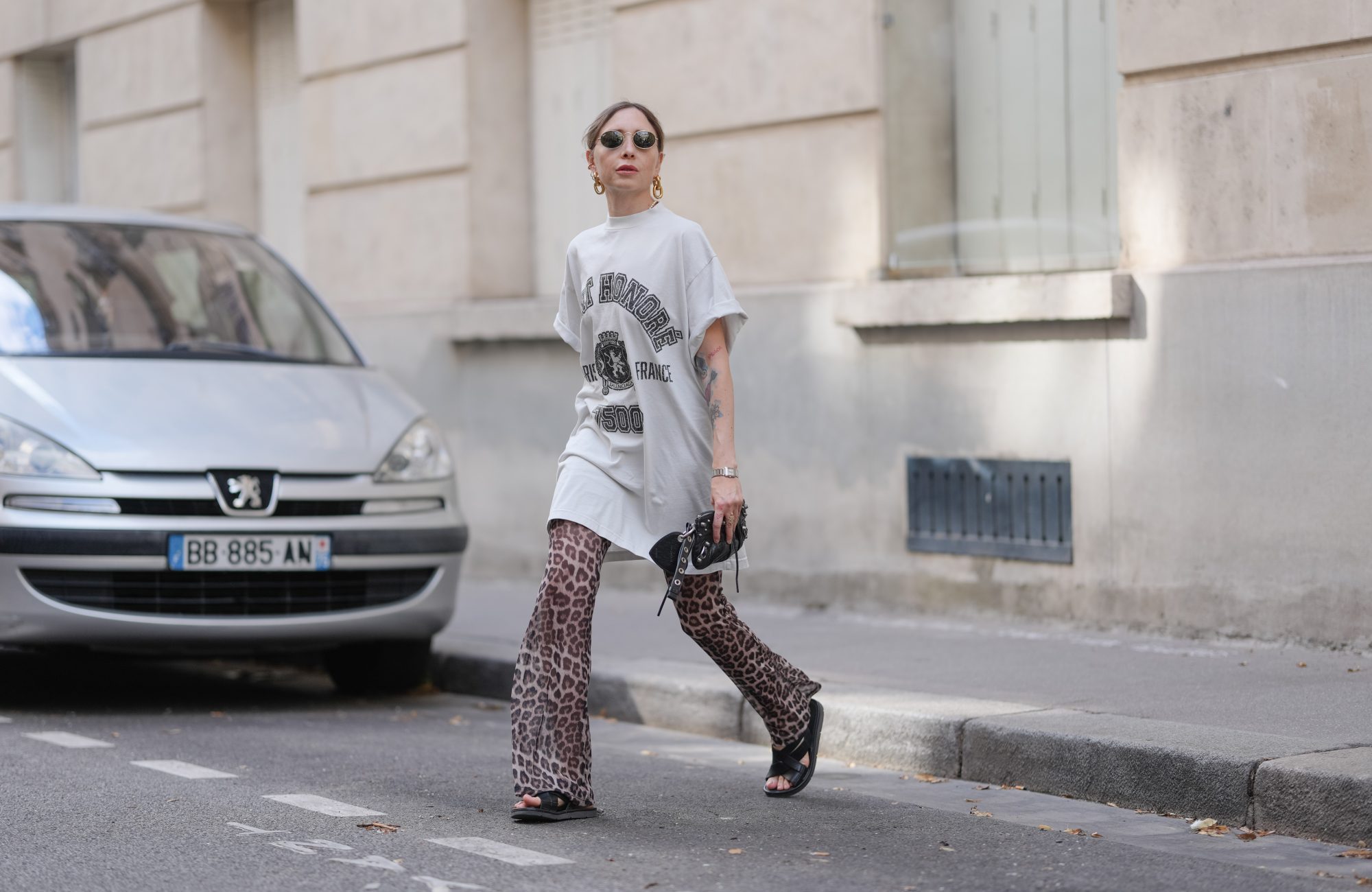 PARIS, FRANCE - JULY 17: Emy Venturini wears an oversized white graphic T-shirt dress by Balenciaga paired with flared leopard print trousers by Calzedonia. Black open-toe flat sandals by Prada complete the look. Accessories include a black leather clutch by Balenciaga, a silver Cartier wristwatch, gold sculptural earrings by Rabanne, and round sunglasses by Ray-Ban. Hair is blond and styled in a low ponytail with a center part, and lips are painted with lipstick in a bold red shade, during a street style fashion photo session, on July 17, 2025 in Paris, France. (Photo by Edward Berthelot/Getty Images)