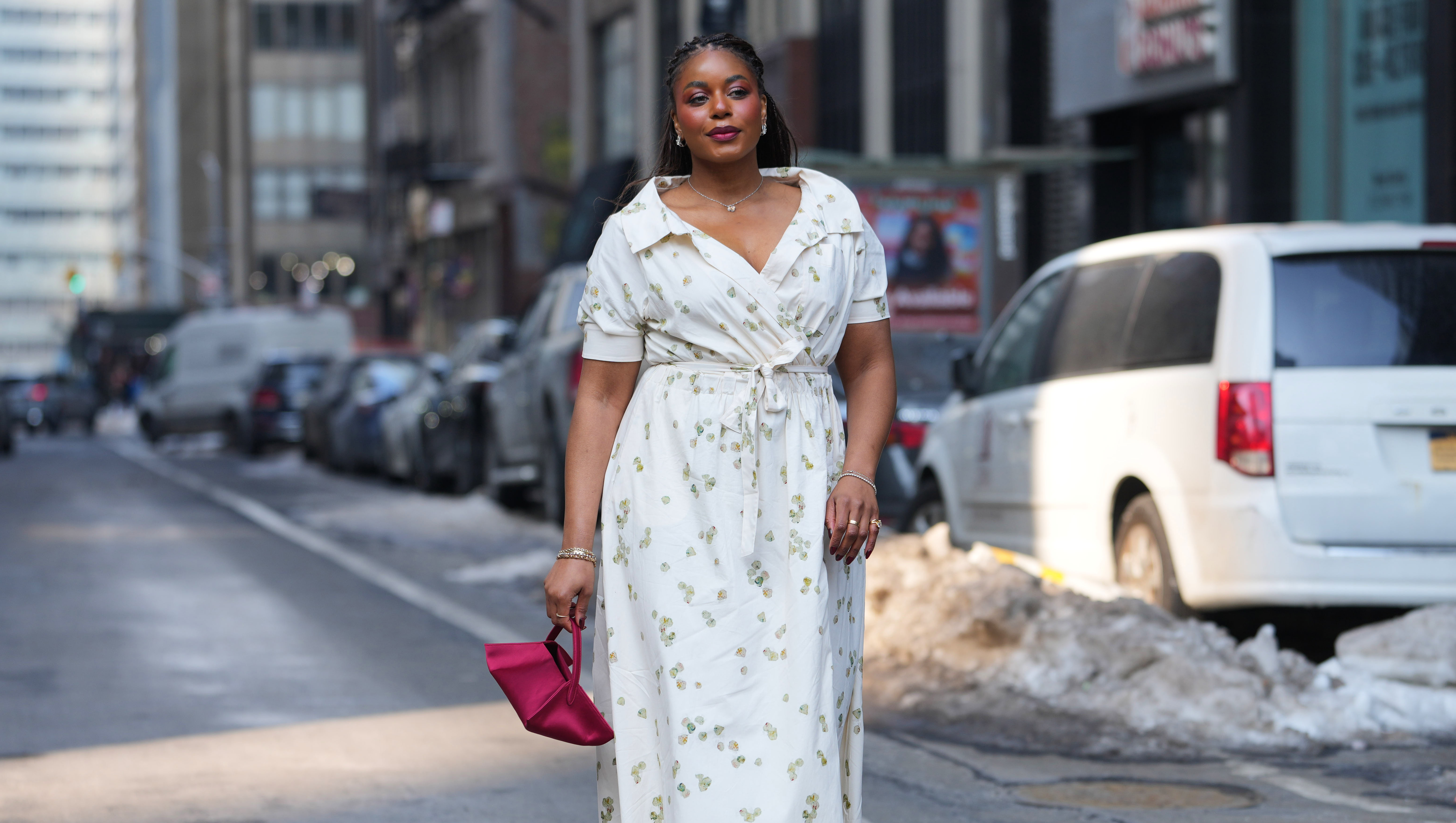 NEW YORK, NEW YORK - FEBRUARY 14: A guest wears braided hair, earrings, necklace, gold rings, a pink satin clutch bag, a white V-neck wrap dress with a tie waist and a floral print, gold metallic open toe sandals shoes, outside Altuzarra, during New York Fashion Week, on February 14, 2026 in New York City (Photo by Edward Berthelot/Getty Images)