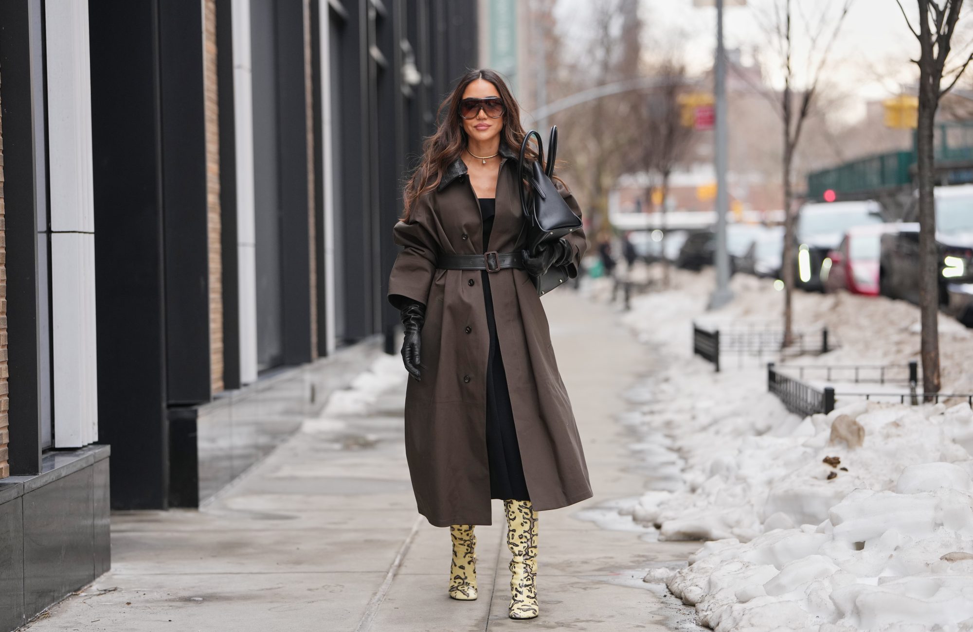 NEW YORK, NEW YORK - FEBRUARY 11: Tamara Kalinic wears oversized black sunglasses, a diamond necklace, a black leather bag, a brown trench coat with a black textured leather collar and a matching belt, a black midi dress, black leather gloves, pale pastel yellow leopard print boots, outside Proenza Schouler, during New York Fashion Week, on February 11, 2026 in New York City (Photo by Edward Berthelot/Getty Images)