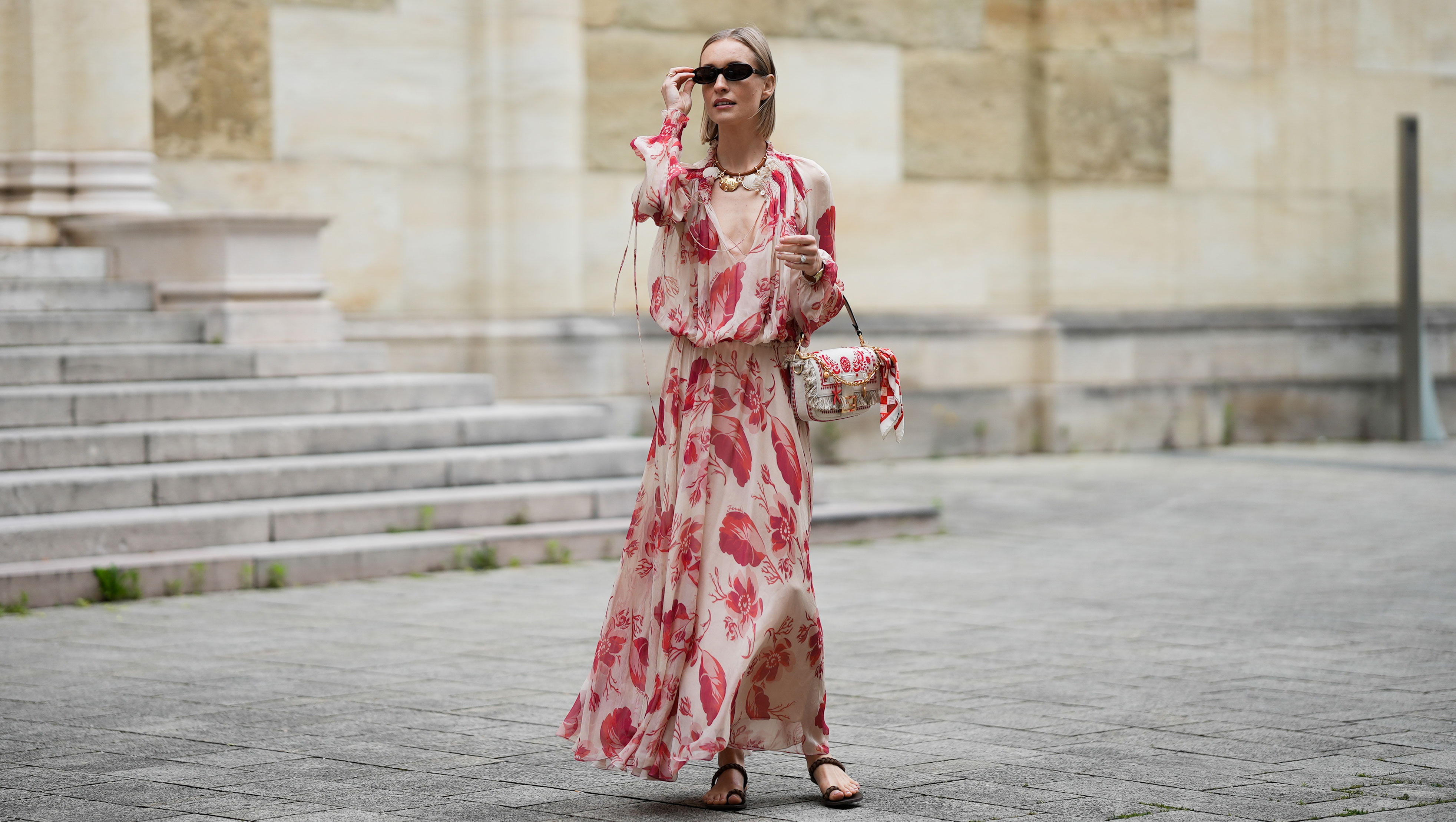 MUNICH, GERMANY - MAY 29: Marlies Pia Pfeifhofer is seen wearing a light beige silk chiffon long sleeve maxi dress with red and pink prints of feathers and flowers, drawstring waist, deep V-neck with front tie and smocking details on the collar and cuffs from FENDI, a natural-colored Baguette canvas bag with coral bandana embroidery, small fringes on the flap, calfskin leather straps, golden hardware and logo detail as well as an additional golden chain metal charm with marine-inspired pendants and a red, pink and white bandana charm with golden logo clip from FENDI, a pair of chocolate brown leather gladiator sandals with a braided leather toe strap and one main strap from Alex Rivière x Manebi, a Louis Tortoise Brown sunglasses with slim and oval shape from Corlin, a gold-colored metal and brown nappa leather Forever Fendi necklace with shell-shaped pendants from FENDI, a brown leather Tank Louis watch with yellow gold rectangular watch case from Cartier, a silver diamond ring on May 29, 2025 in Munich, Germany. (Photo by Moritz Scholz/Getty Images)