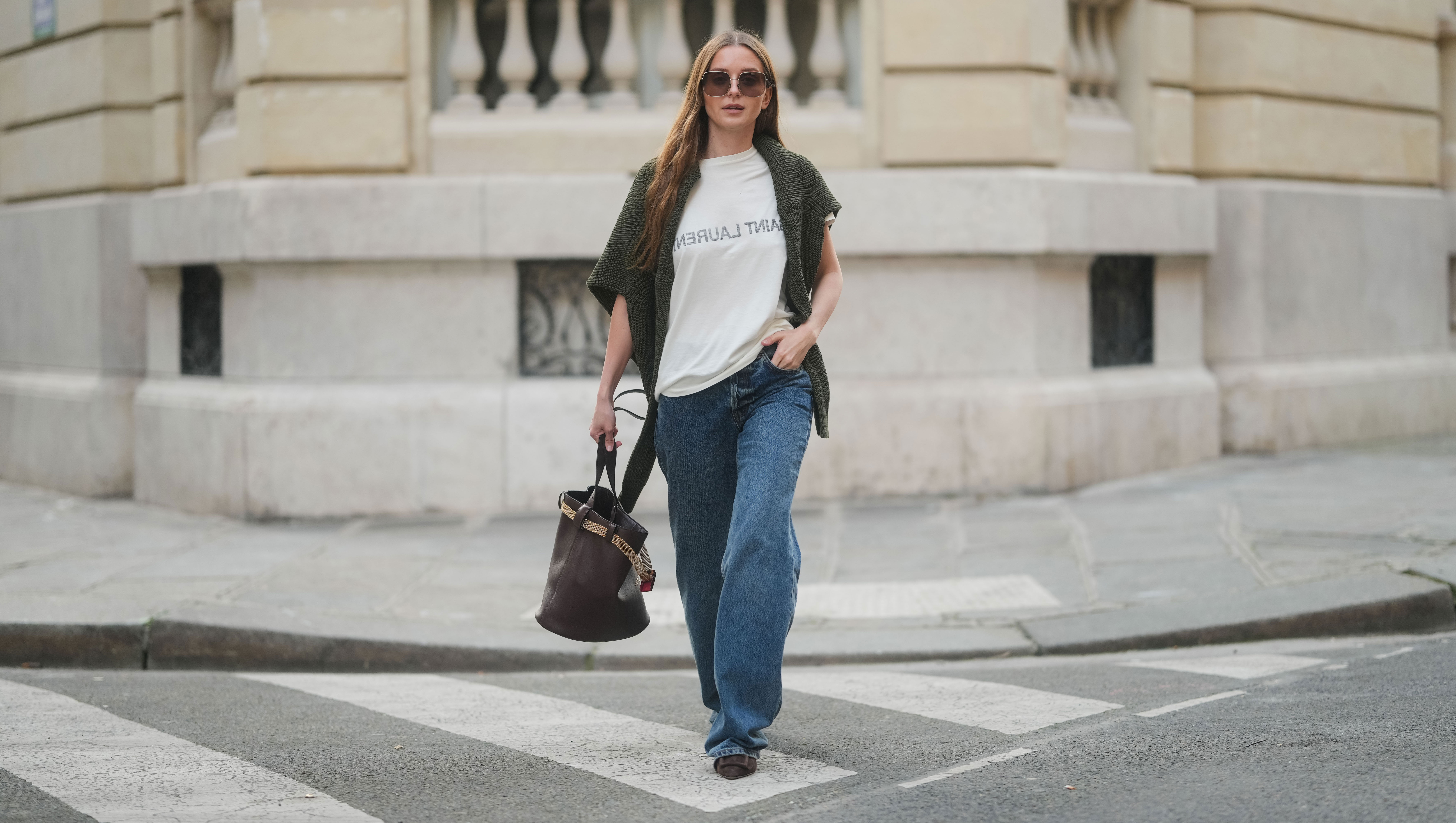 PARIS, FRANCE - MARCH 25: Diane Batoukina wears long straight brown hair with a center part, square Dior sunglasses, a dark green knit sweater by By Aylin Koenig draped over her shoulders, a white Saint Laurent T-shirt, wide-leg blue jeans by Anine Bing, brown suede pointed-toe flats by Manolo Blahnik, and carries a dark brown leather Prada bucket bag, during a street style fashion photo session on March 25, 2025 in Paris, France. (Photo by Edward Berthelot/Getty Images)