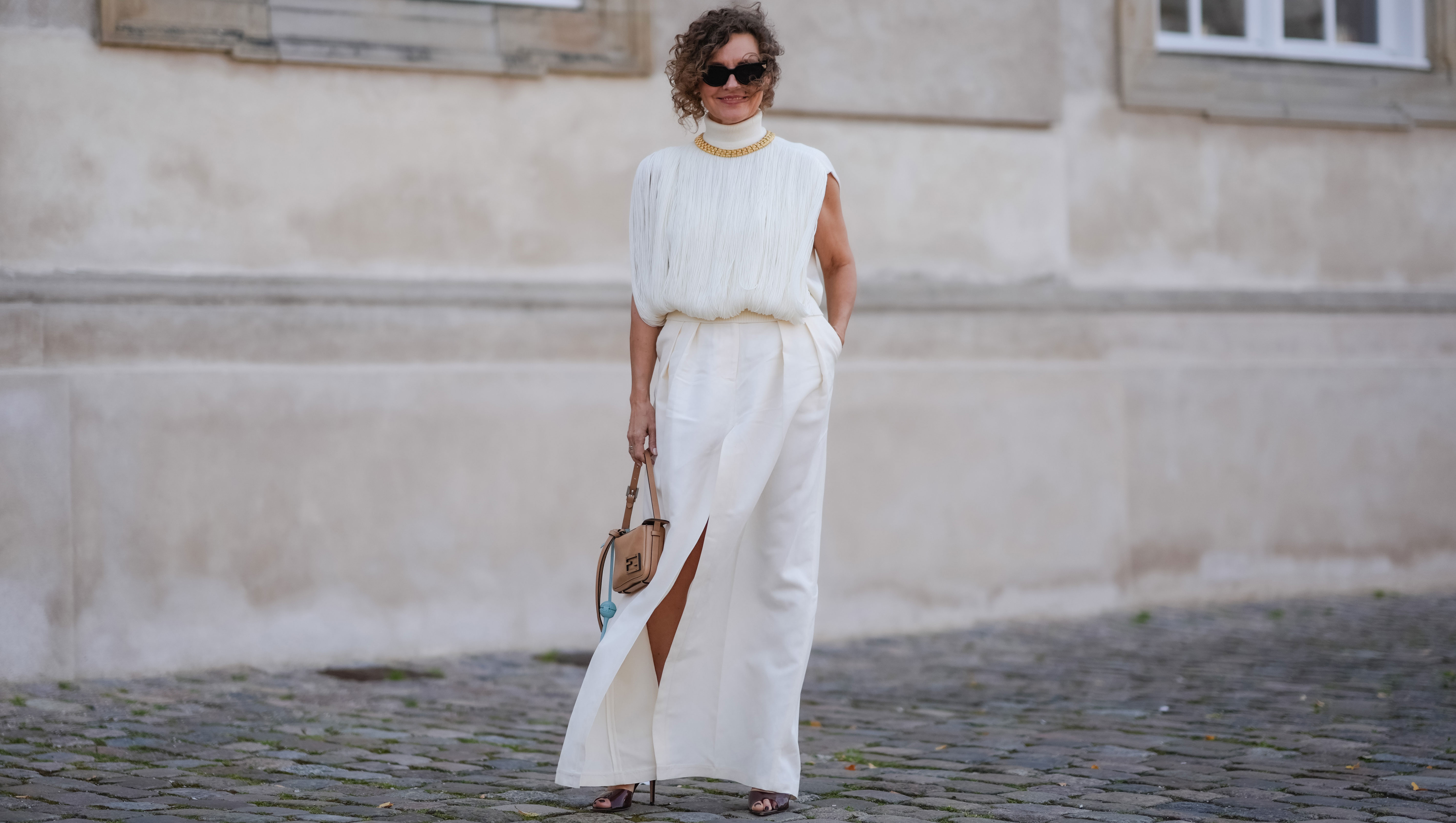 COPENHAGEN, DENMARK - AUGUST 7: Renia Jaz wears black sunglasses, gold necklace, white high neck fringe sleeveless shirt, white maxi skirt with high slit, light brown bag, light blue Fendi lollipop leather holder, dark brown mule heels, outside The Garment, during the Copenhagen Fashion Week Spring/Summer 2024-2025 on August 7, 2024 in Copenhagen, Denmark. (Photo by Edward Berthelot/Getty Images)