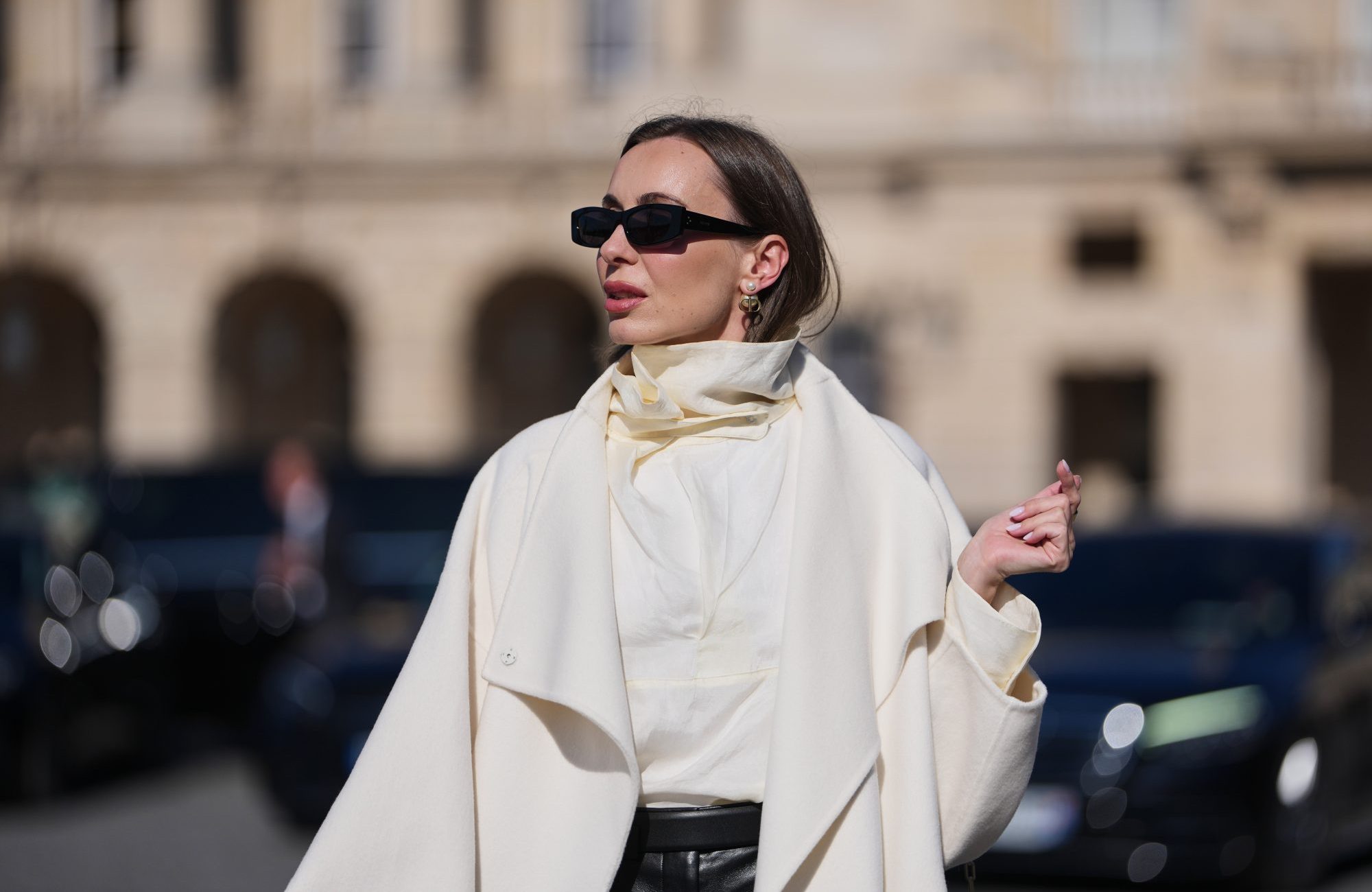 PARIS, FRANCE - MARCH 03: A guest wears black squared sunglasses, a gold logo and white pearls pendant earrings from Dior, a white wool long coat, black leather micro shorts, a white silk high neck blouse, outside Dior, during Paris Fashion Week - Womenswear Fall/Winter 2026/2027, on March 03, 2026 in Paris, France (Photo by Edward Berthelot/Getty Images)