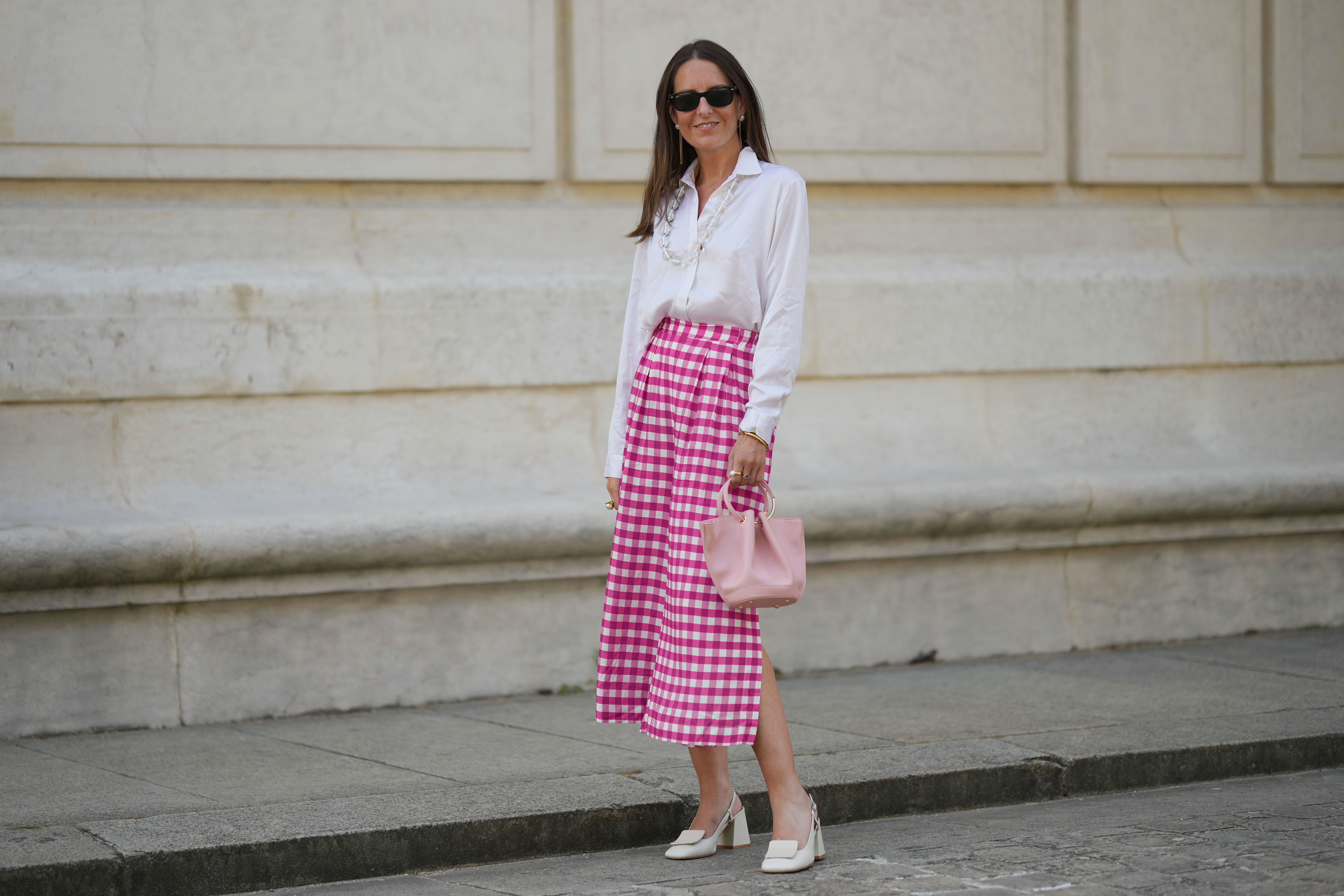 Alba Garavito Torre wears black sunglasses from Ray Ban, white pearl and gold chain pendant earrings, a transparent large pearls long necklace, a white shirt, pink and white checkered print pattern midi pants from Frambuã, gold bracelets, a pale pink matte leather handbag from Saonara Brand, white latte matte shiny leather block heels mules from Jonak, during a street style fashion photo session, on June 06, 2023 in Paris, France.