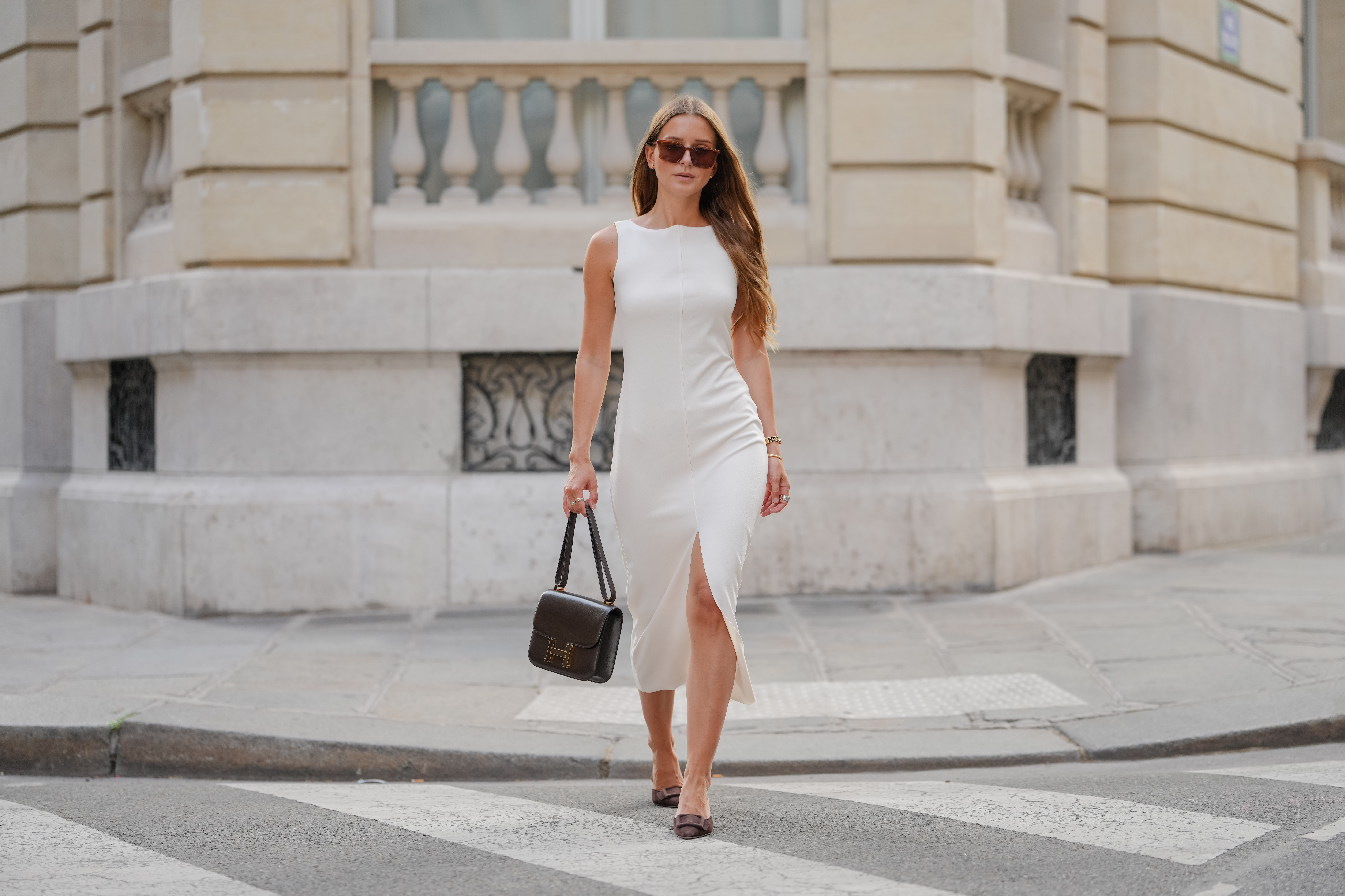 Diane Batoukina wears a sleeveless, fitted white dress with a high neckline and a midi length. Long, loose hair is styled with a center part. Sunglasses feature a brown tint and a rectangular shape. A black leather handbag from Hermes with a structured design and a prominent gold "H" detail is carried. On the wrist, a delicate gold bracelet is visible. Footwear consists of pointed-toe shoes in a dark color, during a street style fashion photo session, on June 13, 2025 in Paris, France