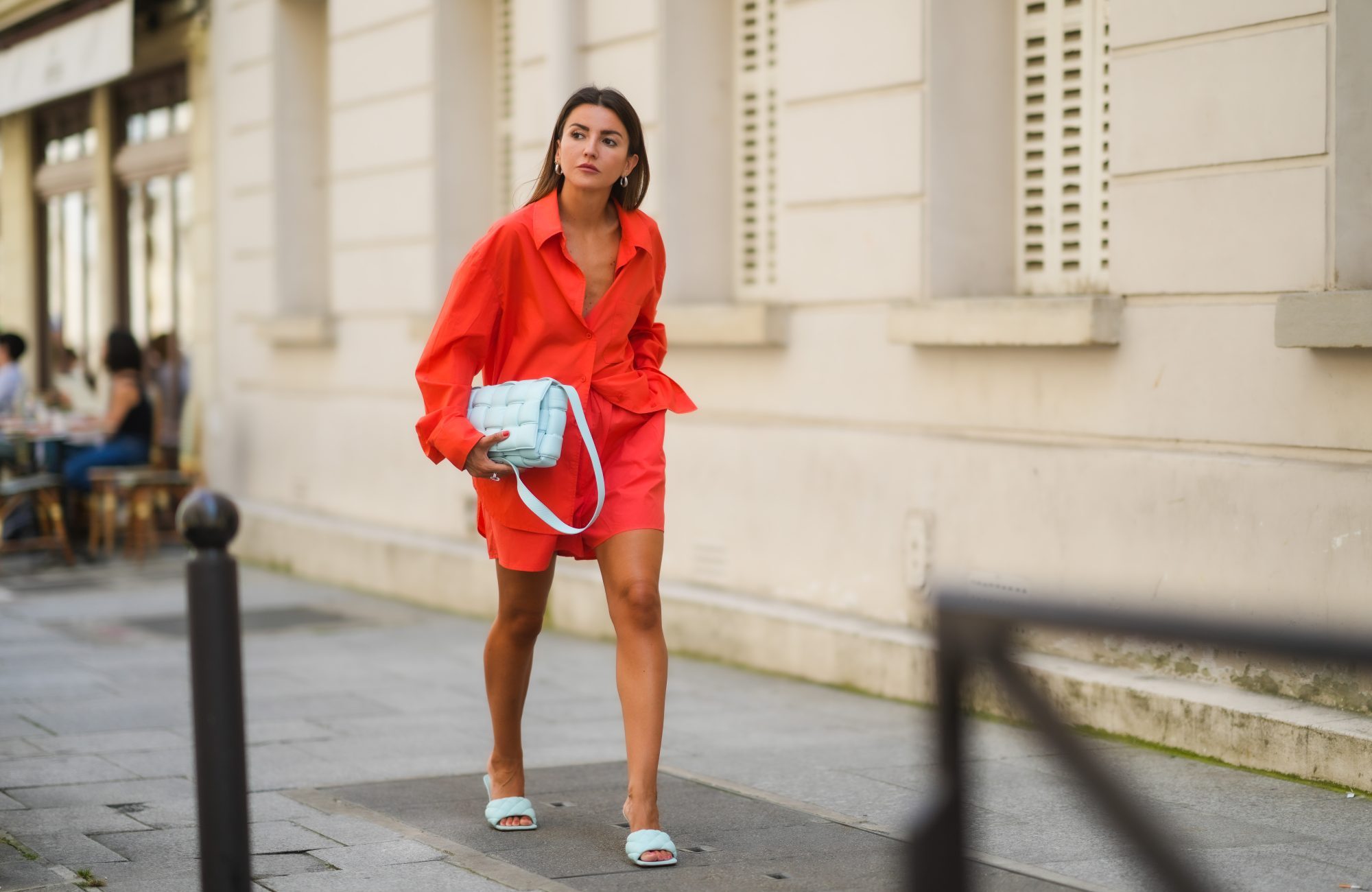 Alexandra Pereira wears earrings, a bold neon orange / red oversized shirt, matching mini shorts, a pale pastel blue woven leather bag from Bottega Veneta, pale pastel quilted leather sandals from Bottega Veneta, on July 20, 2021 in Paris, France.
