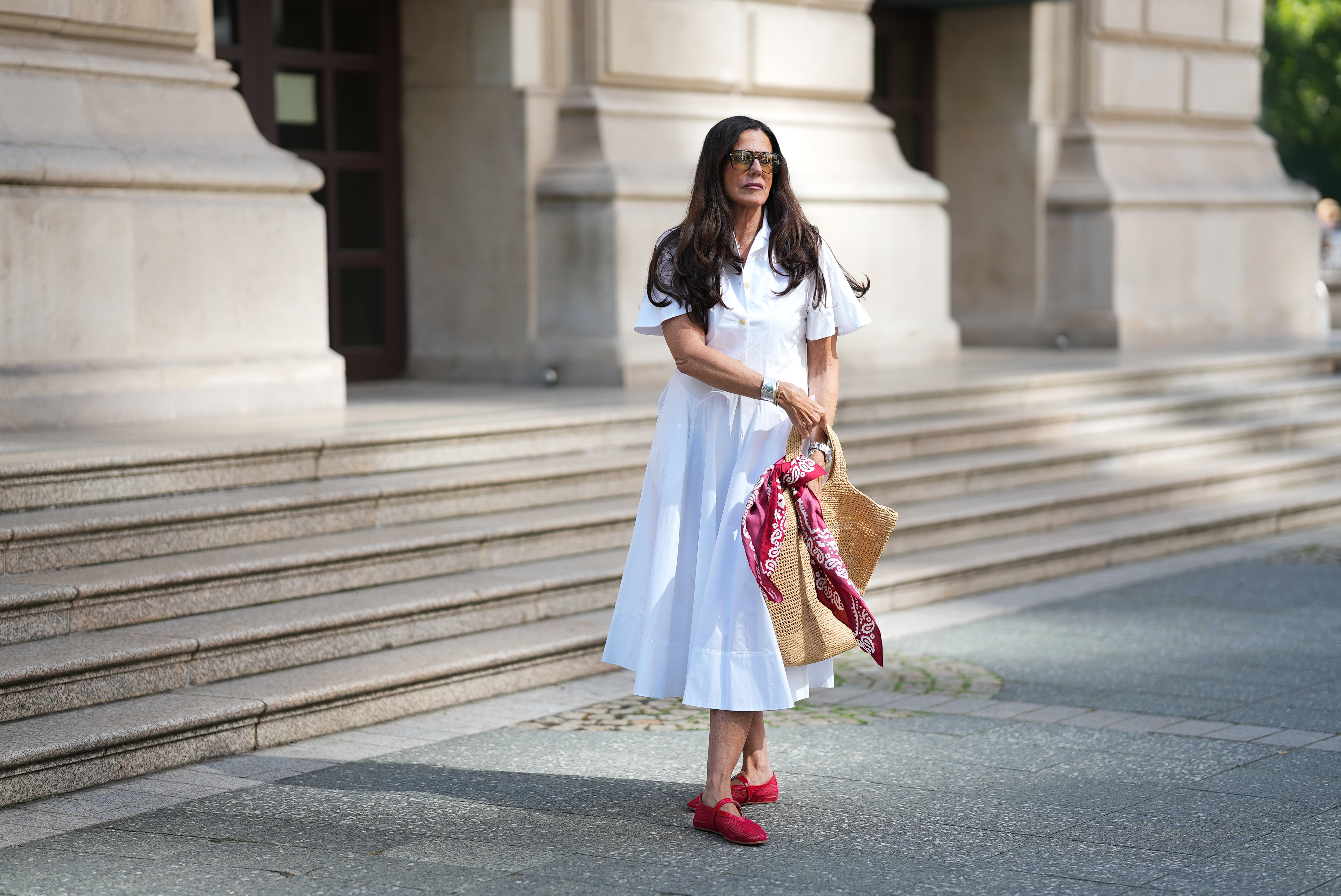 Angelika Hanel is seen wearing a white short-sleeved shirt midi dress with a flared silhouette and soft pleating from Erika Cavallini; a pair of signature Ala&iuml;a ballet flats in red fishnet textile and patent leather, a buckle strap on top and round toes from Ala&iuml;a; the ICARE maxi shopper crafted from hand-embroidered natural raffia and a brown gold oversized, sculptured Cassandre from Saint Laurent adorned with a red and white silk bandana from Arket tied to the handle; a silver Rolex Daytona watch; a yellow golden Juste un Clou bracelet from Cartier; a pav&eacute; diamond Tennis bracelet; a bold sterling silver Bone Cuff from Tiffany & Co.; oversized black aviator sunglasses with orange shaded glasses from Kapten & Son; long brown hair and natural make-up on July 10, 2025 in Frankfurt am Main, Germany.
