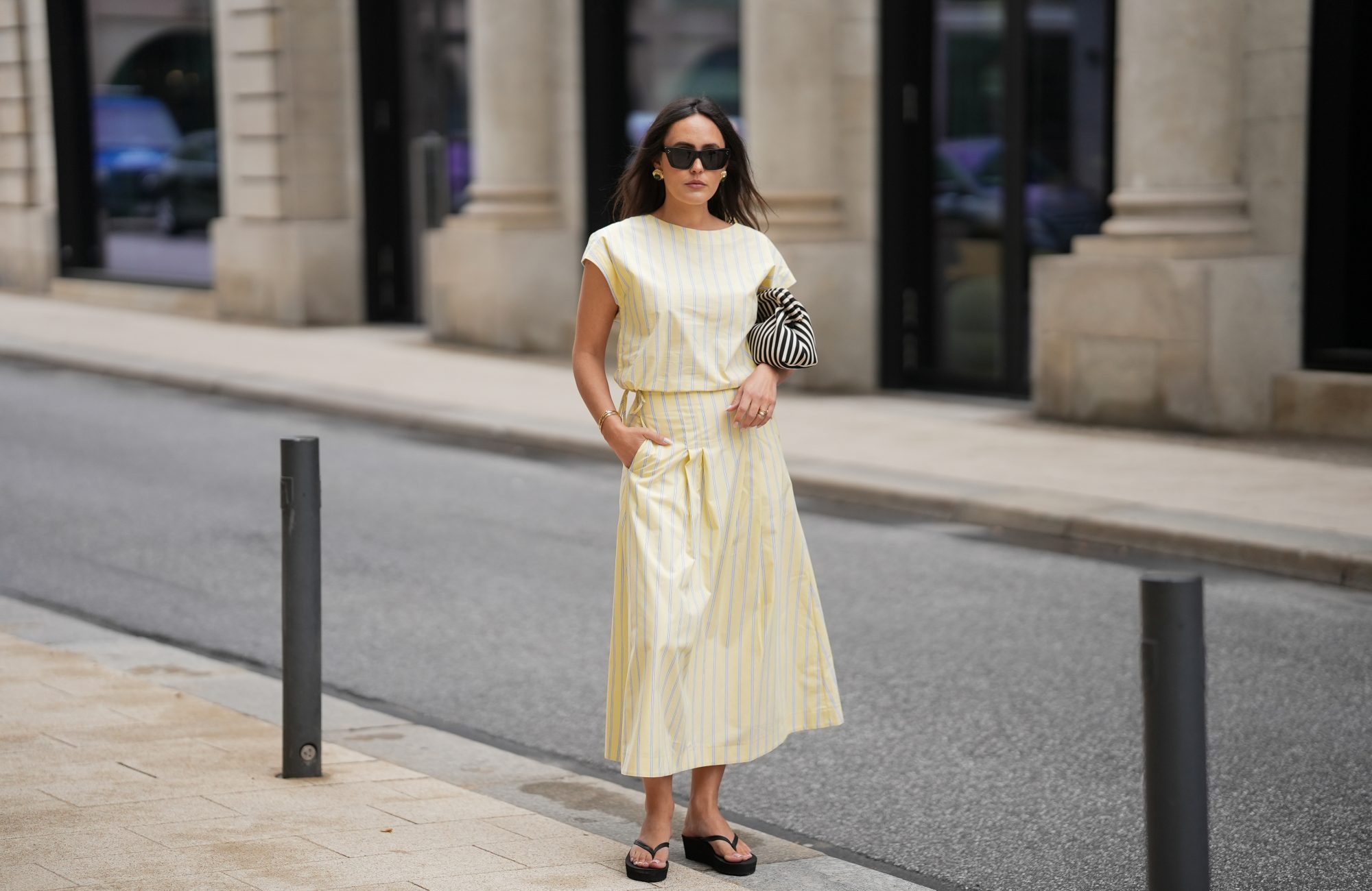 Laura Noltemeyer is seen wearing a yellow striped two-piece set with a sleeveless draped top and matching midi skirt by Sézane; black flip-flop sandals by Amazon; a black-and-white striped Porter clutch bag; black cat-eye sunglasses by Saint Laurent; nude-colored fingernails and toenails; gold jewelry by H&M and COS consisting of earrings, bangles and rings; natural makeup; her dark brown hair is worn in soft waves with a middle part on May 5, 2026 in Hamburg, Germany.