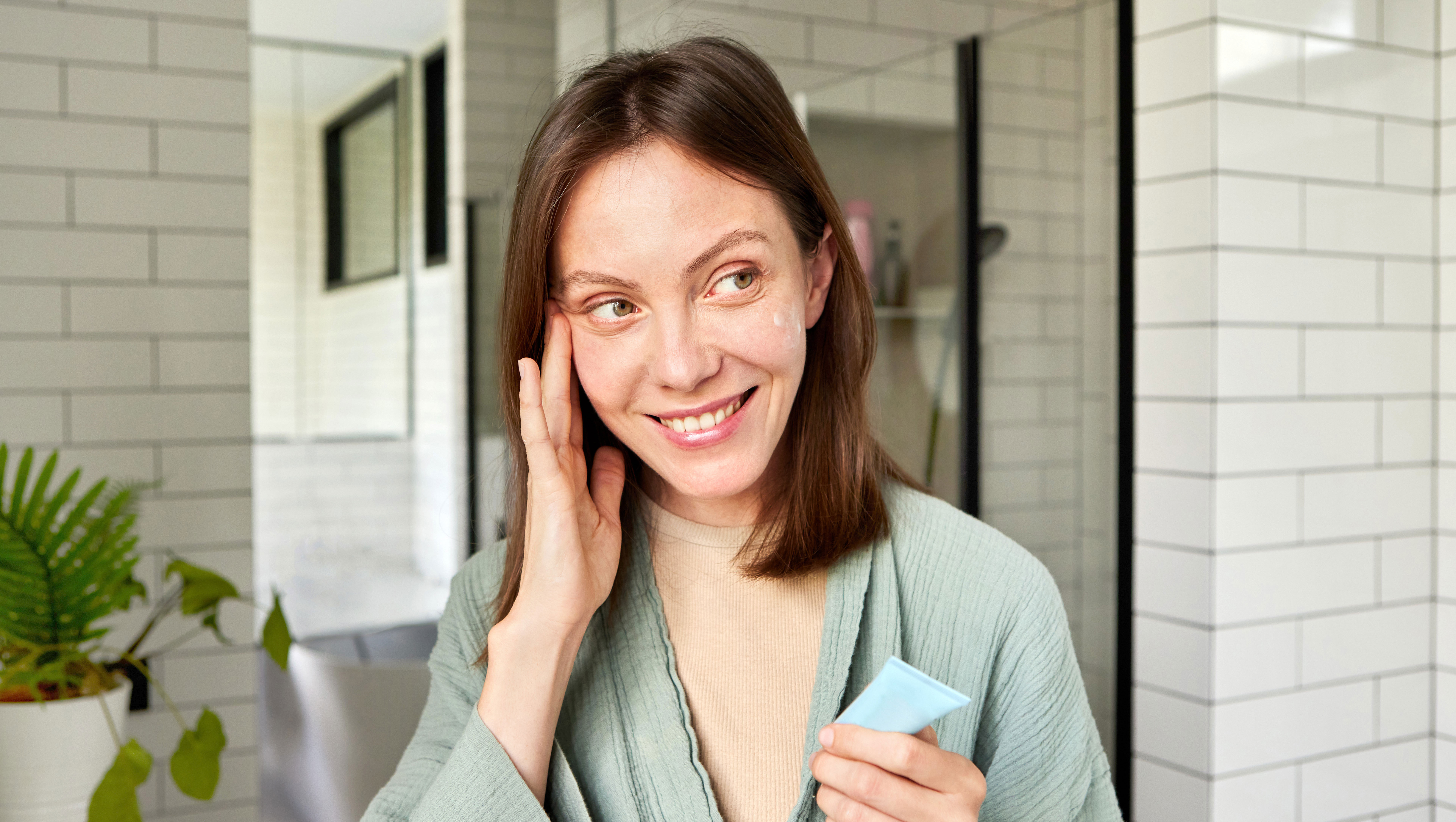 Smiling young woman in a bathrobe applying moisturizing cream to her face after a refreshing shower in a modern bathroom, enjoying her daily self-care routine and promoting healthy, glowing skin