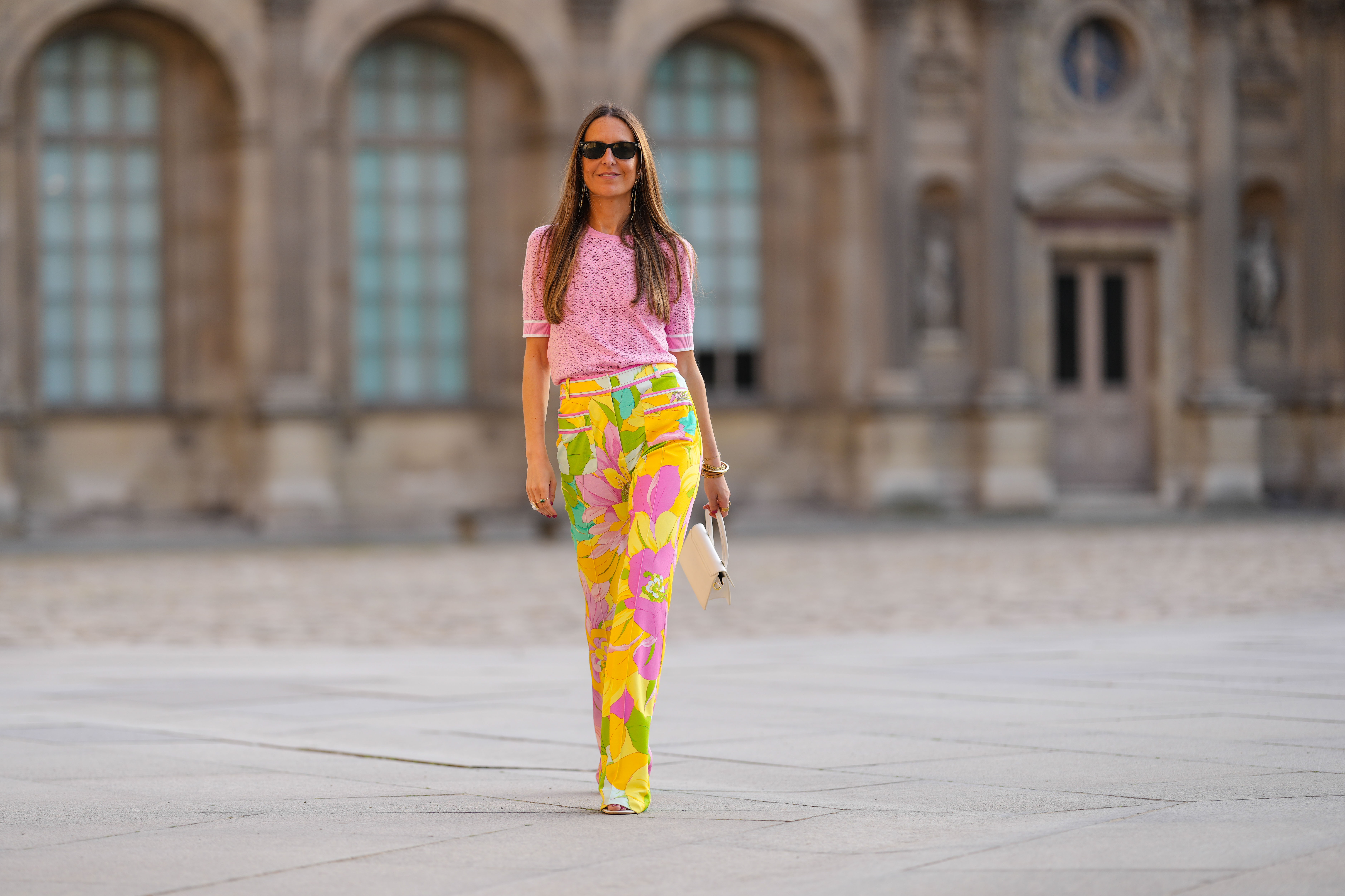 Alba Garavito Torre wears black sunglasses from Ray Ban, gold earrings, a pale pink cut-out pattern short sleeves t-shirt Leonard Paris, high waist yellow / green / pink flower print pattern flared pants, white latte matte leather Chiquito handbag from Jacquemus, a pink gold Love bracelet from Cartier, gold bracelets, gold rings, white leather Oasis block heels sandals from Hermes, during a street style fashion photo session, on April 22, 2022 in Paris, France.