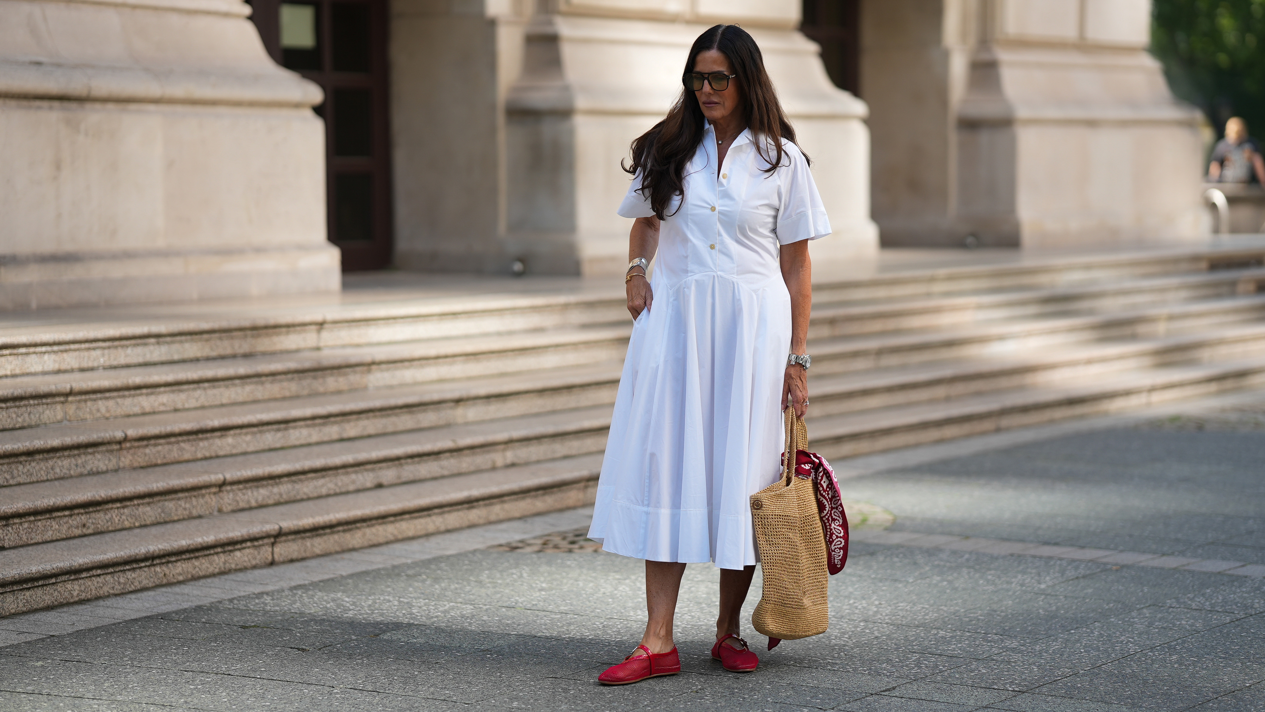 FRANKFURT AM MAIN, GERMANY - JULY 10: Angelika Hanel is seen wearing a white short-sleeved shirt midi dress with a flared silhouette and soft pleating from Erika Cavallini; a pair of signature Ala&iuml;a ballet flats in red fishnet textile and patent leather, a buckle strap on top and round toes from Ala&iuml;a; the ICARE maxi shopper crafted from hand-embroidered natural raffia and a brown gold oversized, sculptured Cassandre from Saint Laurent adorned with a red and white silk bandana from Arket tied to the handle; a silver Rolex Daytona watch; a silver diamond ring; a yellow golden Juste un Clou bracelet from Cartier; a pav&eacute; diamond Tennis bracelet; a bold sterling silver Bone Cuff from Tiffany & Co.; oversized black aviator sunglasses with orange shaded glasses from Kapten & Son; long brown hair and natural make-up on July 10, 2025 in Frankfurt am Main, Germany. (Photo by Moritz Scholz/Getty Images)