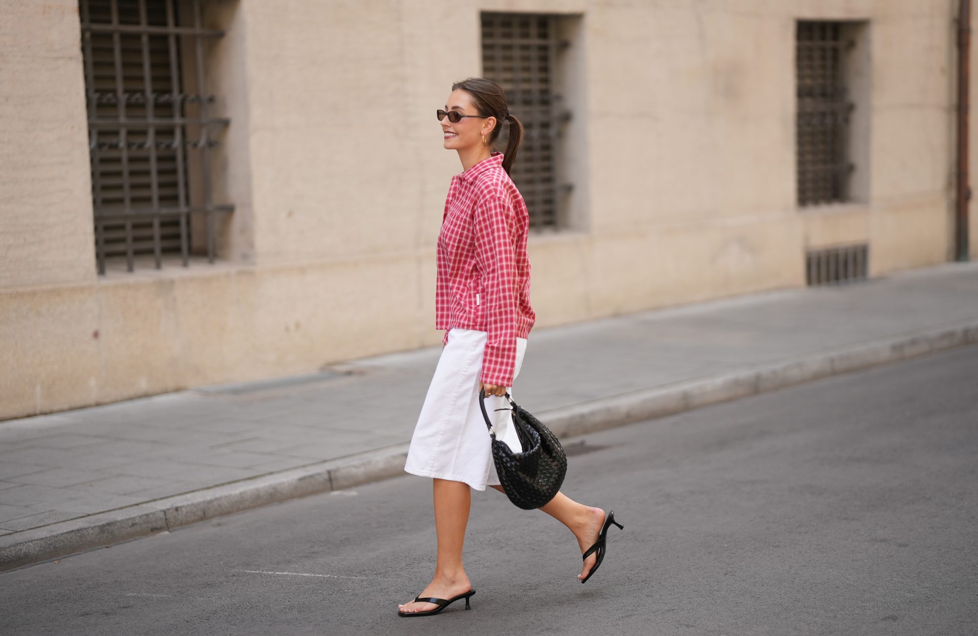 MUNICH, GERMANY - JUNE 8: Lina Uhrig is seen wearing a pink and red cotton checked cropped shirt with a button front closure, long sleeves, pointed collar, loose fit silhouette and small check pattern from JJXX; white cotton Bermuda shorts with a straight silhouette, knee length, clean waistband and minimal design from Stradivarius; black leather kitten heel sandals with thin toe straps, open toe construction and low slim heels from Stradivarius; a natural pedicure visible through the sandals; a black woven leather shoulder bag with a compact curved silhouette, short top handles, textured woven surface and metal hardware details from Adax; gold tone hoop earrings; rectangular black acetate sunglasses with narrow frames and dark tinted lenses from Chimi; her brown hair is worn in a sleek low ponytail with a center part; with natural make up on June 8, 2026 in Munich, Germany. (Photo by Moritz Scholz/Getty Images)