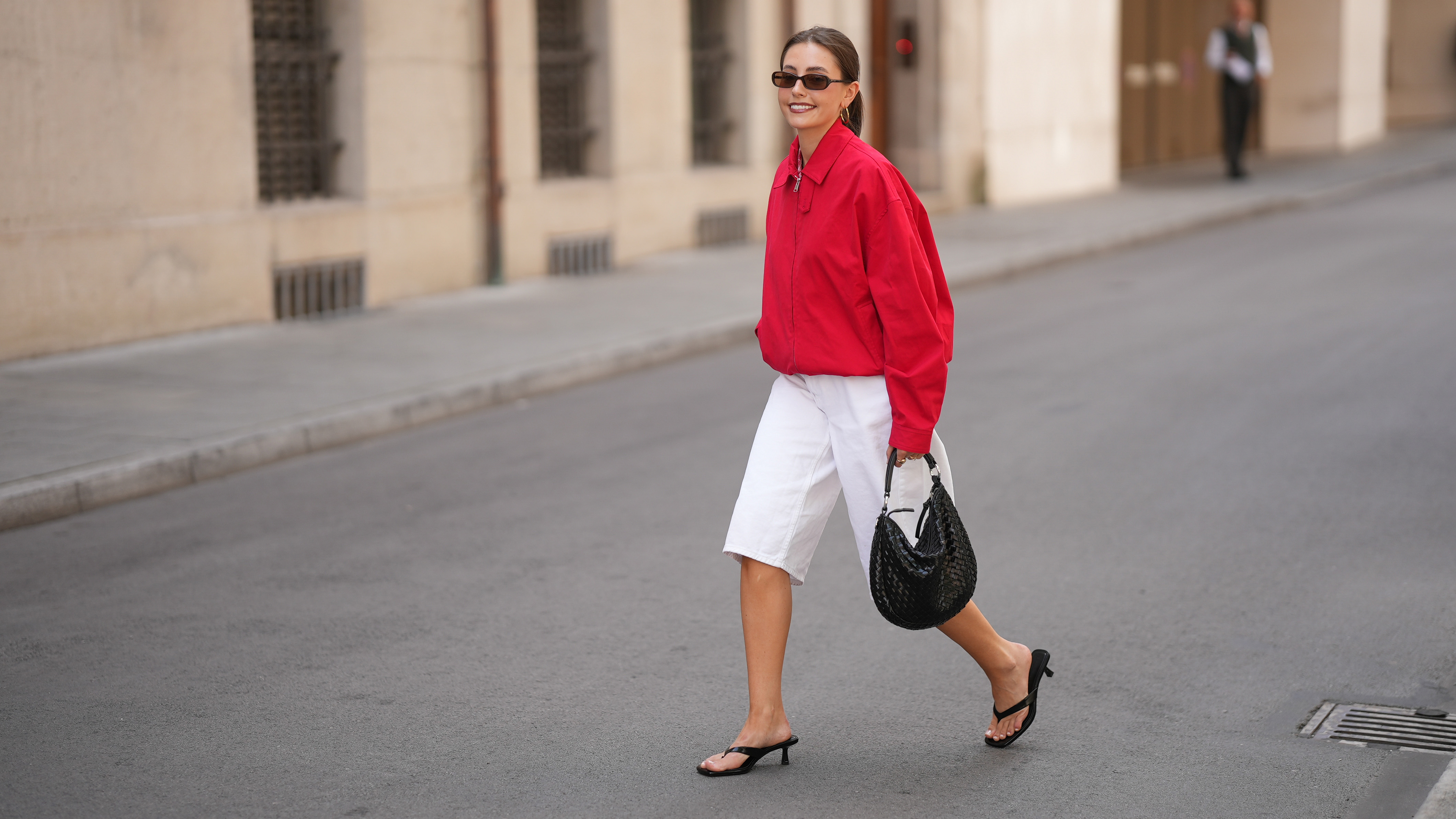 MUNICH, GERMANY - JUNE 8: Lina Uhrig is seen wearing a red cotton cropped jacket with a shirt collar, front zip closure, long sleeves, relaxed oversized silhouette and minimal design from Mango x Hailey Bieber; white cotton Bermuda shorts with a straight silhouette, knee length, clean waistband and minimal design from Stradivarius; black leather kitten heel sandals with thin toe straps, open toe construction and low slim heels from Stradivarius; a natural pedicure visible through the sandals; a black woven leather shoulder bag with a compact curved silhouette, short top handles, textured woven surface and metal hardware details from Adax; gold tone hoop earrings; rectangular black acetate sunglasses with narrow frames and dark tinted lenses from Chimi; her brown hair is worn in a sleek low ponytail with a center part; with natural make up on June 8, 2026 in Munich, Germany. (Photo by Moritz Scholz/Getty Images)